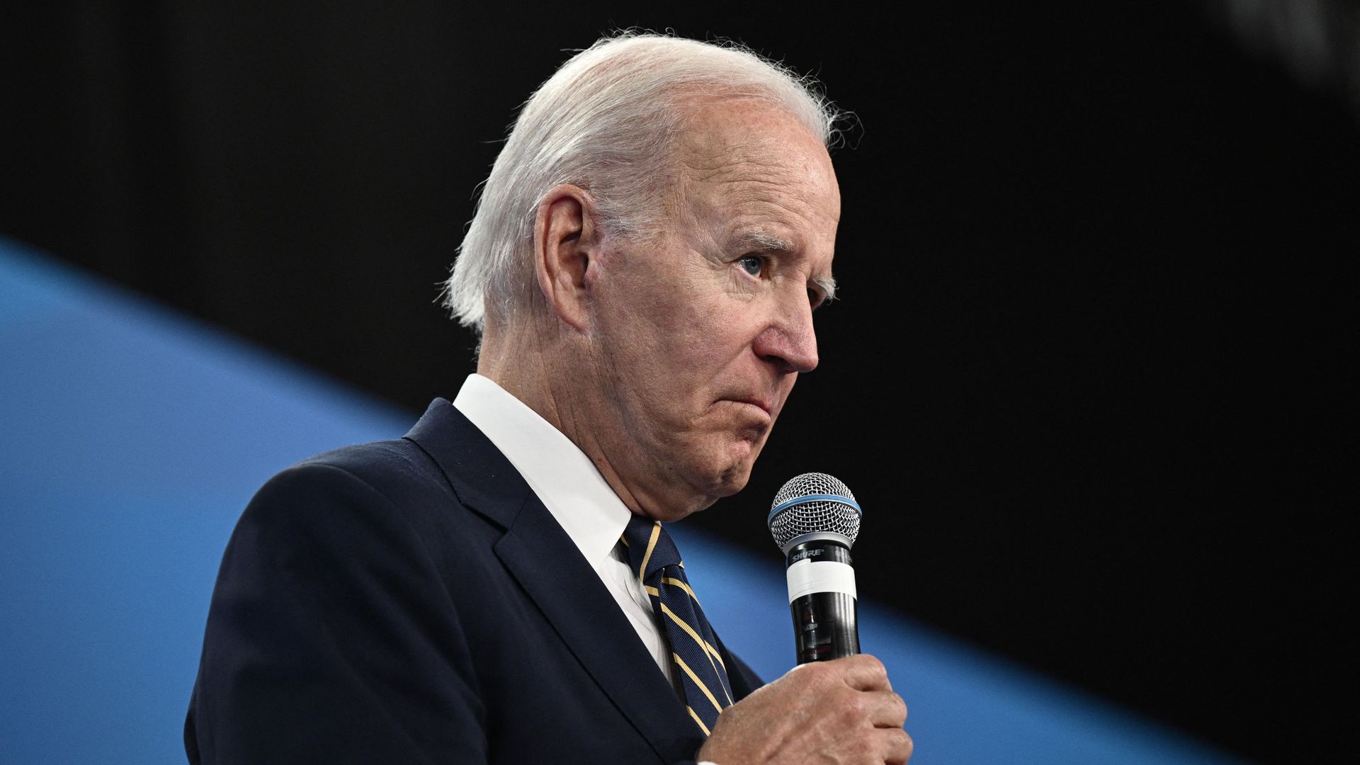 President Joe Biden holds a microphone, and the background is black and blue.