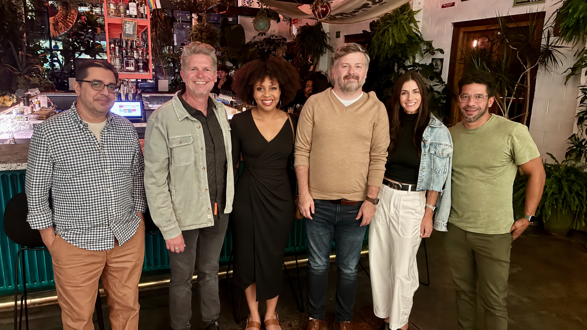 Six people pose in a bar, smiling at the camera; a diverse group in casual outfits stands in a row, with bottles, plants, and warm lighting behind them.