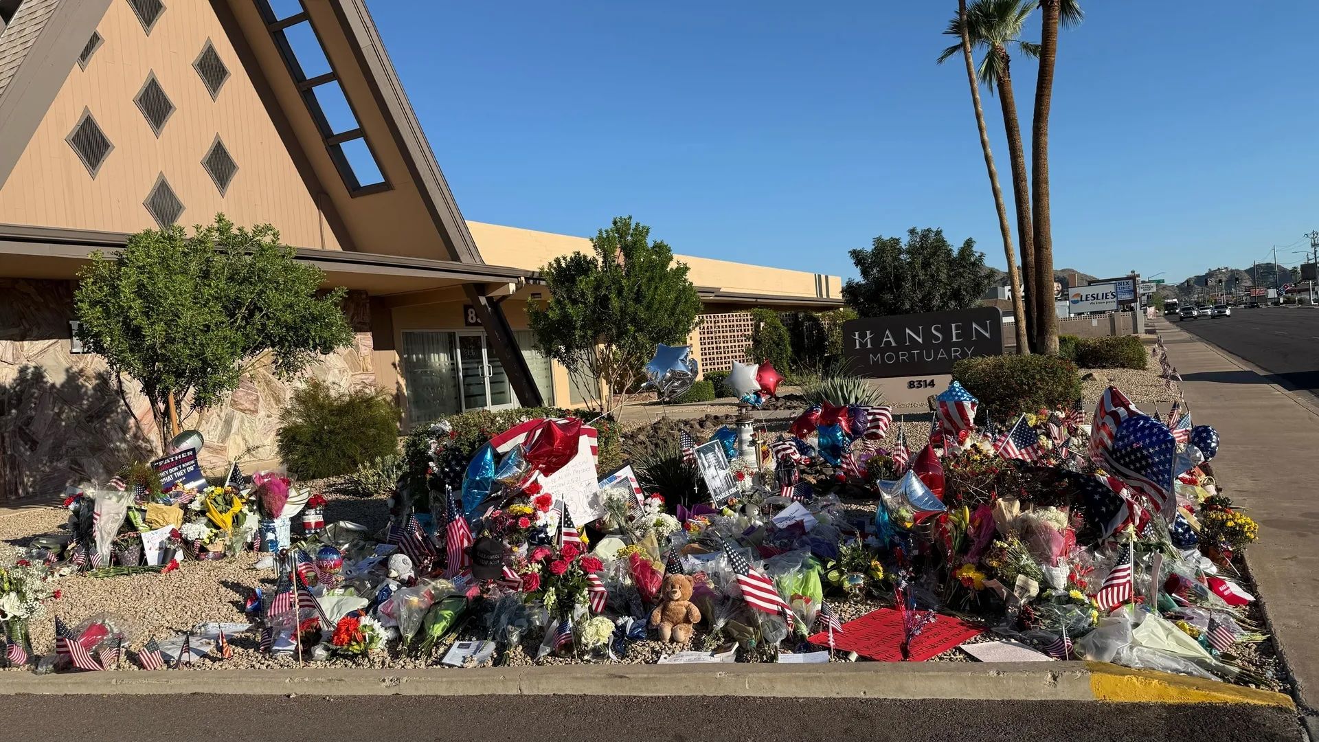Charlie Kirk memorial outside Hansen Mortuary with numerous flowers, American flags, balloons in red, white, and blue, and a teddy bear under a clear blue sky near palm trees.