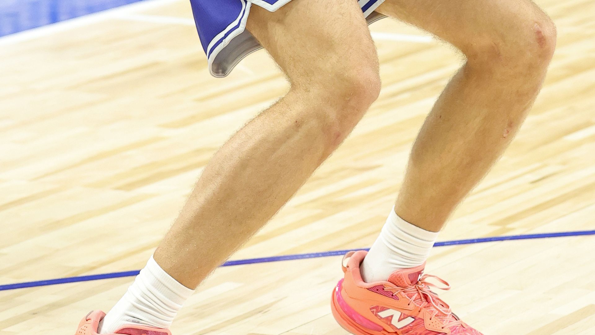 A person wearing blue basketball shorts with a draft combine logo and bright pink New Balance sneakers is standing on a basketball court. The focus is on the legs and shoes against the hardwood floor.