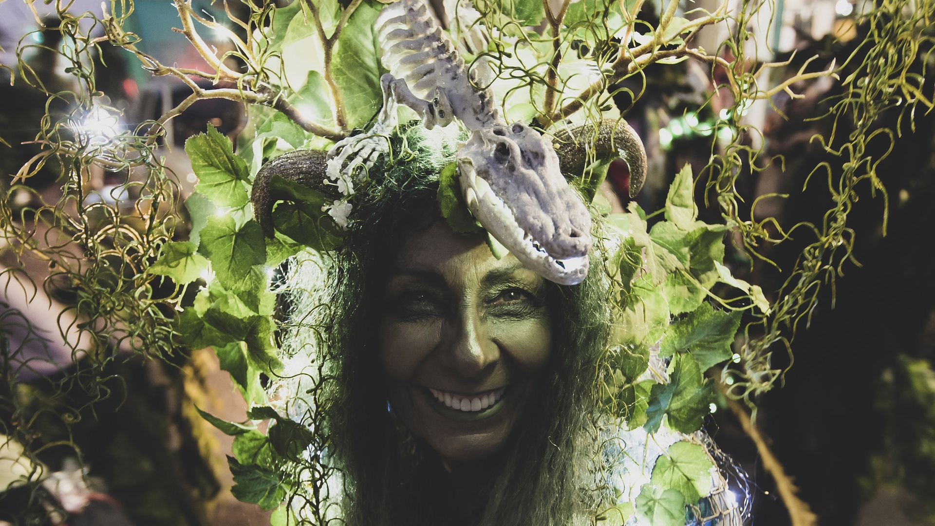 Smiling person wearing a costume with green leaves, twisted vines, horns, and a crocodile skull headpiece, illuminated by small white lights against a dark background.