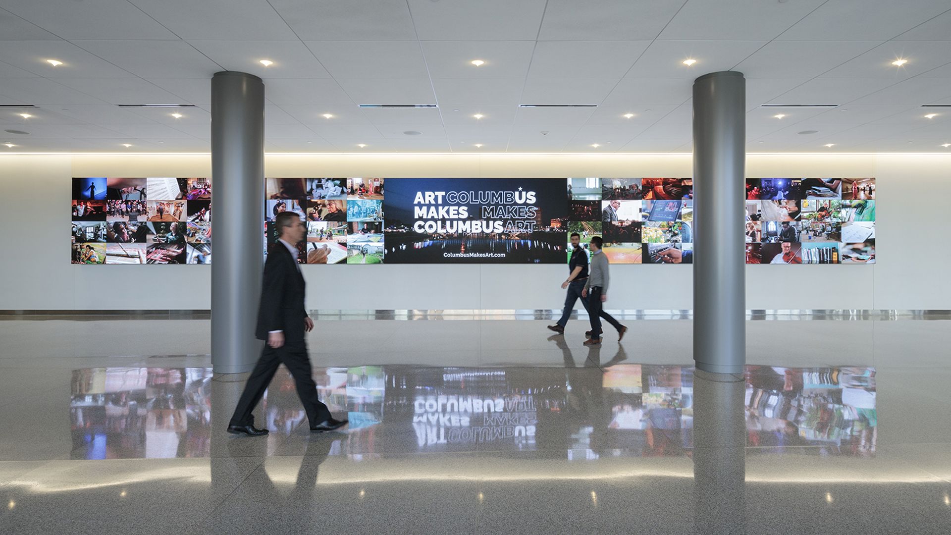 Spacious modern atrium with two large gray columns and a collage wall. A central black panel reads "ART COLUMBUS MAKES COLUMBUS ART" flanked by colorful photo tiles; two people walk on a glossy, reflective floor.