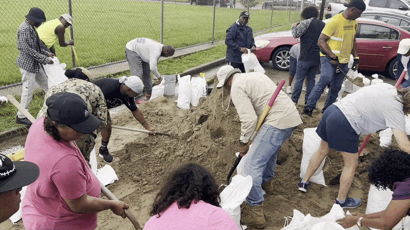About a dozen people fill and tie off sandbags from a central pile of sand.