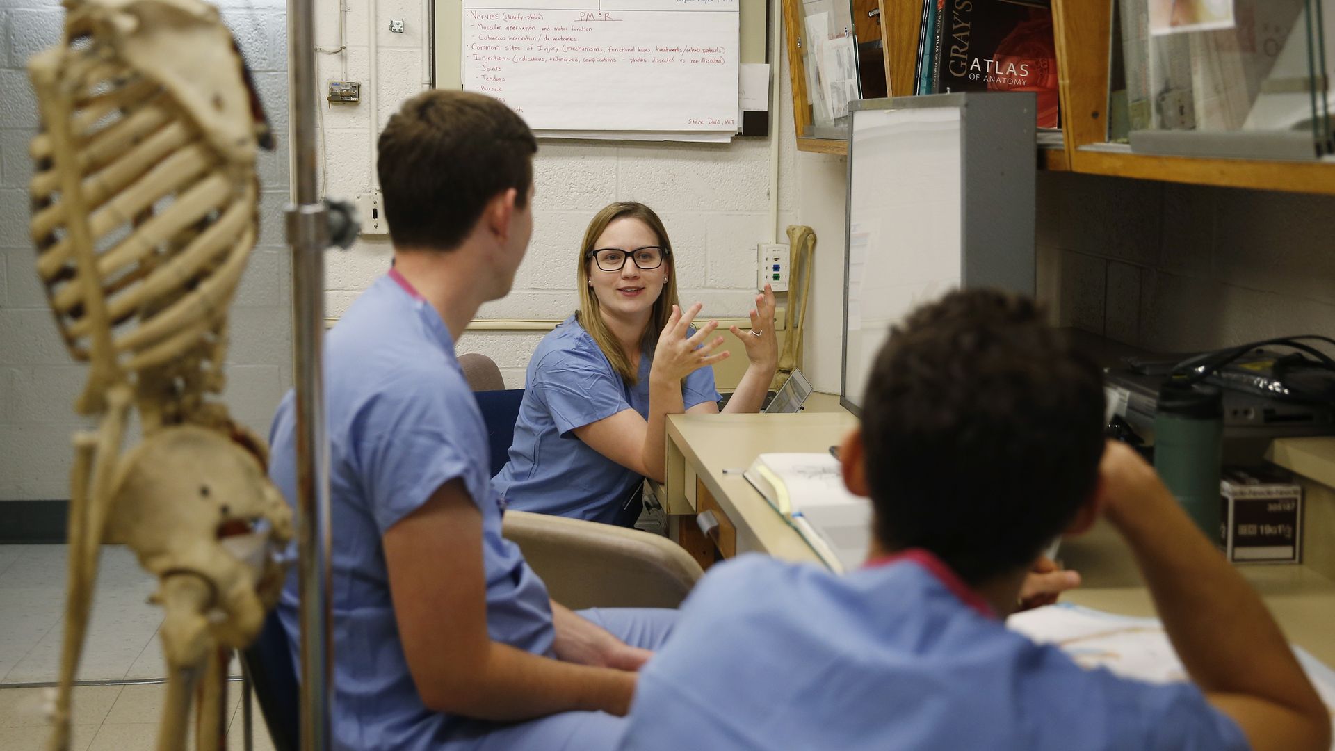 Medical students sit in an office, talking, next to a skeleton.