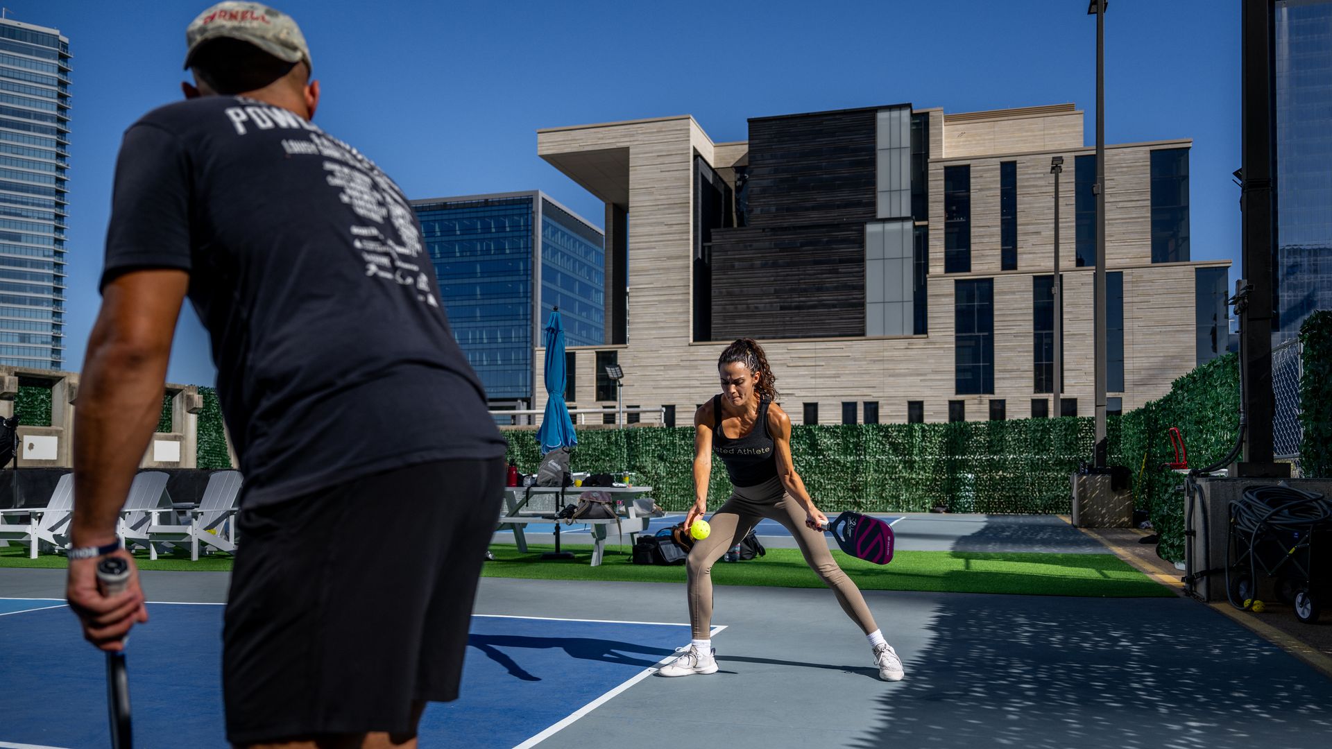 Outdoor padel court with a clear blue sky. A man in a cap stands left; an athletic woman crouches near the net with a paddle and ball, modern beige buildings in the background.