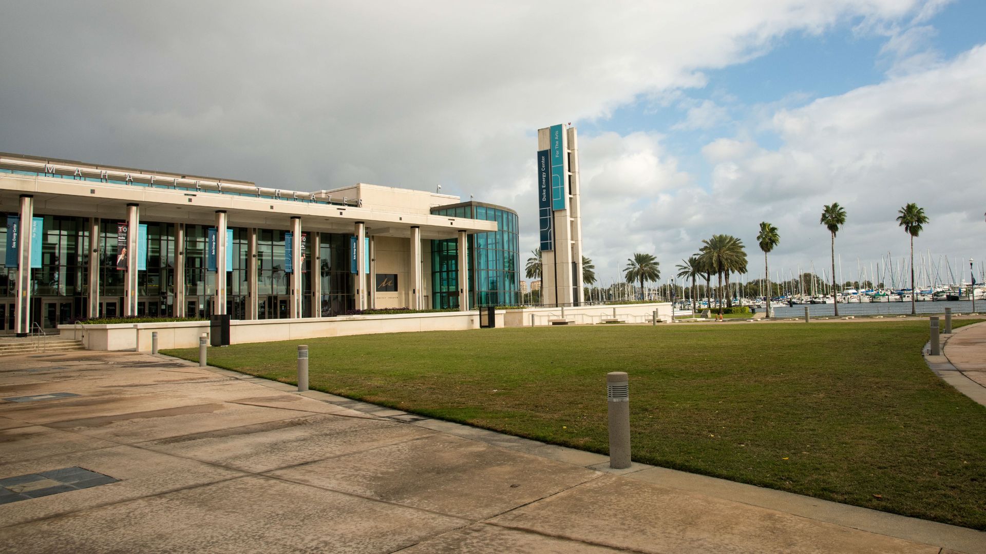 Modern waterfront building with glass walls and turquoise banners sits along a lawn. A tall sign tower overlooks a marina of sailboats, with palm trees and a cloudy blue sky.