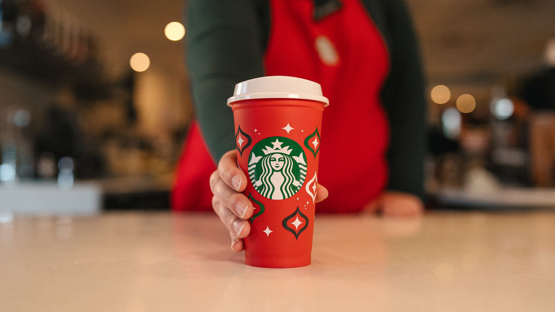 Starbucks red reusable cup with white lid in forefront being held by employee wearing red apron and black long sleeve shirt.