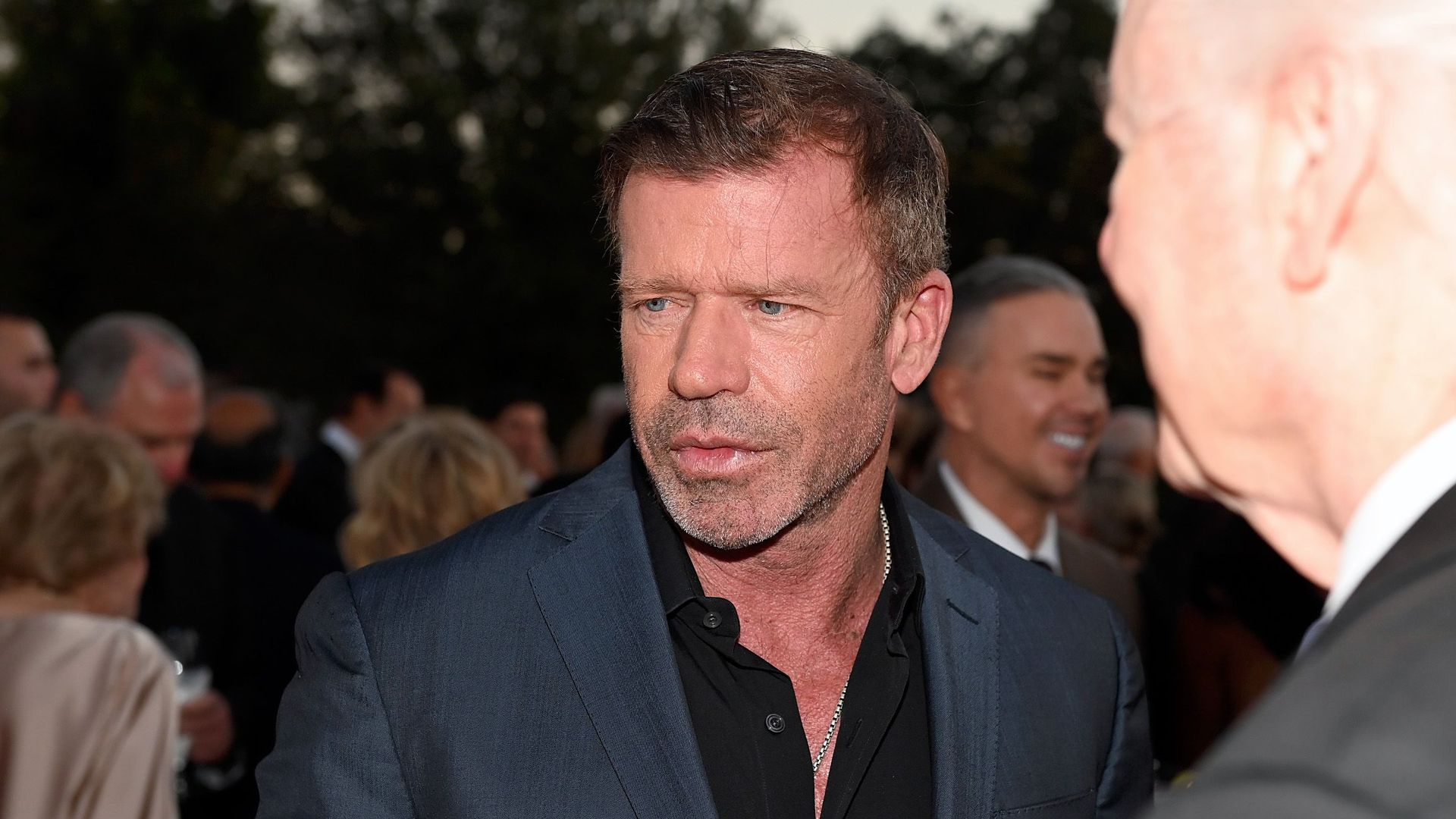 Taylor Sheridan in Dallas in October is seen wearing a black button-down shirt and a navy blue blazer and looking toward the left of the frame.