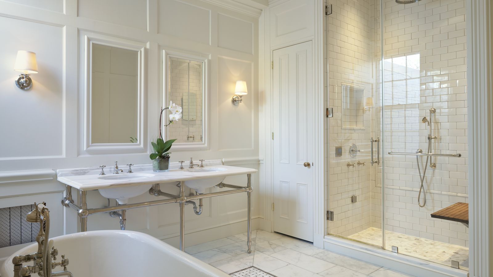 A photo showing a white bathroom with two sinks, exposed plumbing underneath, a see-through shower with glass doors and multiple sprayers and a bathtub