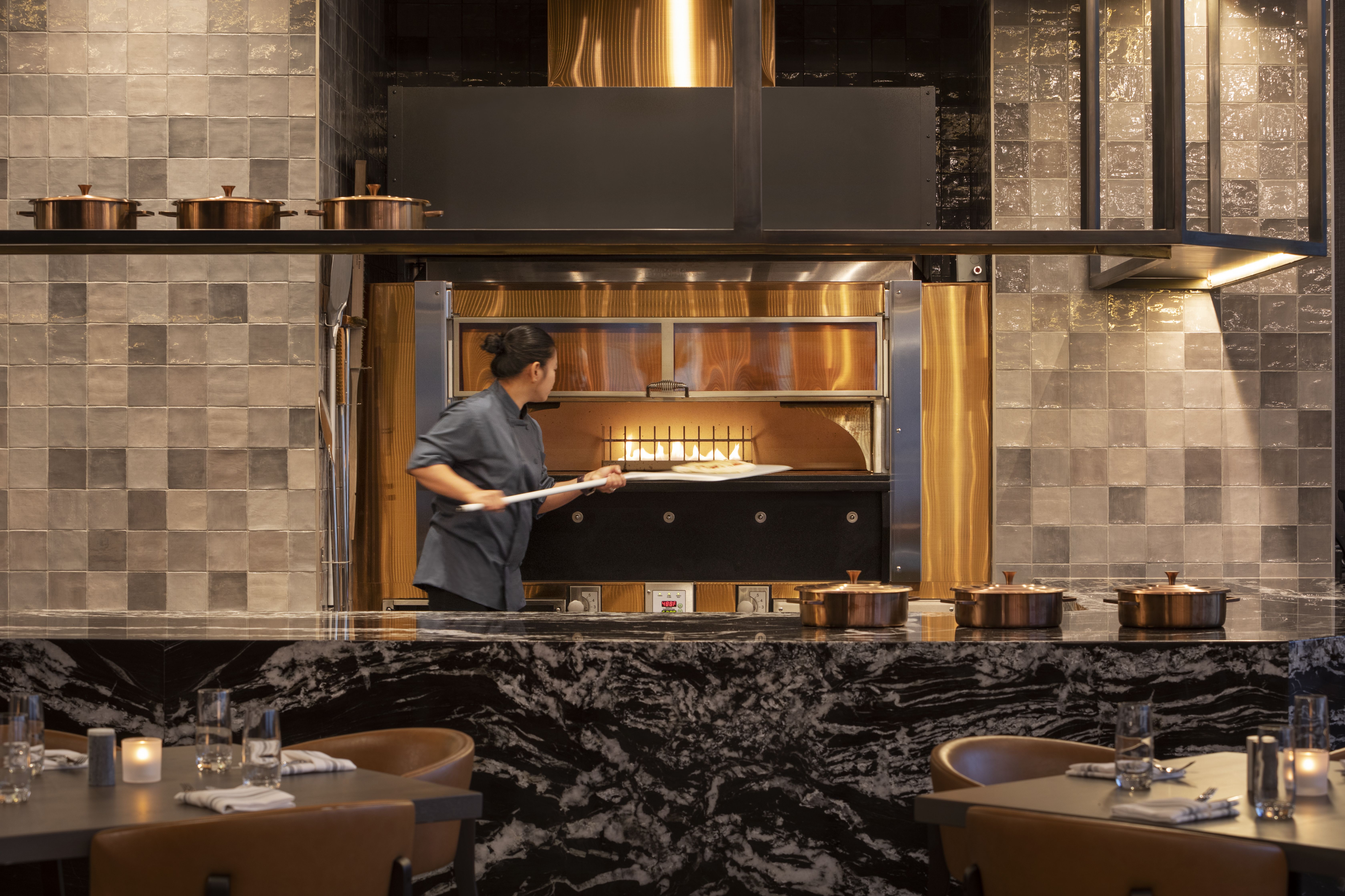 Chef in dark uniform placing pizza into wood-fired oven with flames, in modern restaurant kitchen with copper pots on marble counter and gray tiled walls.