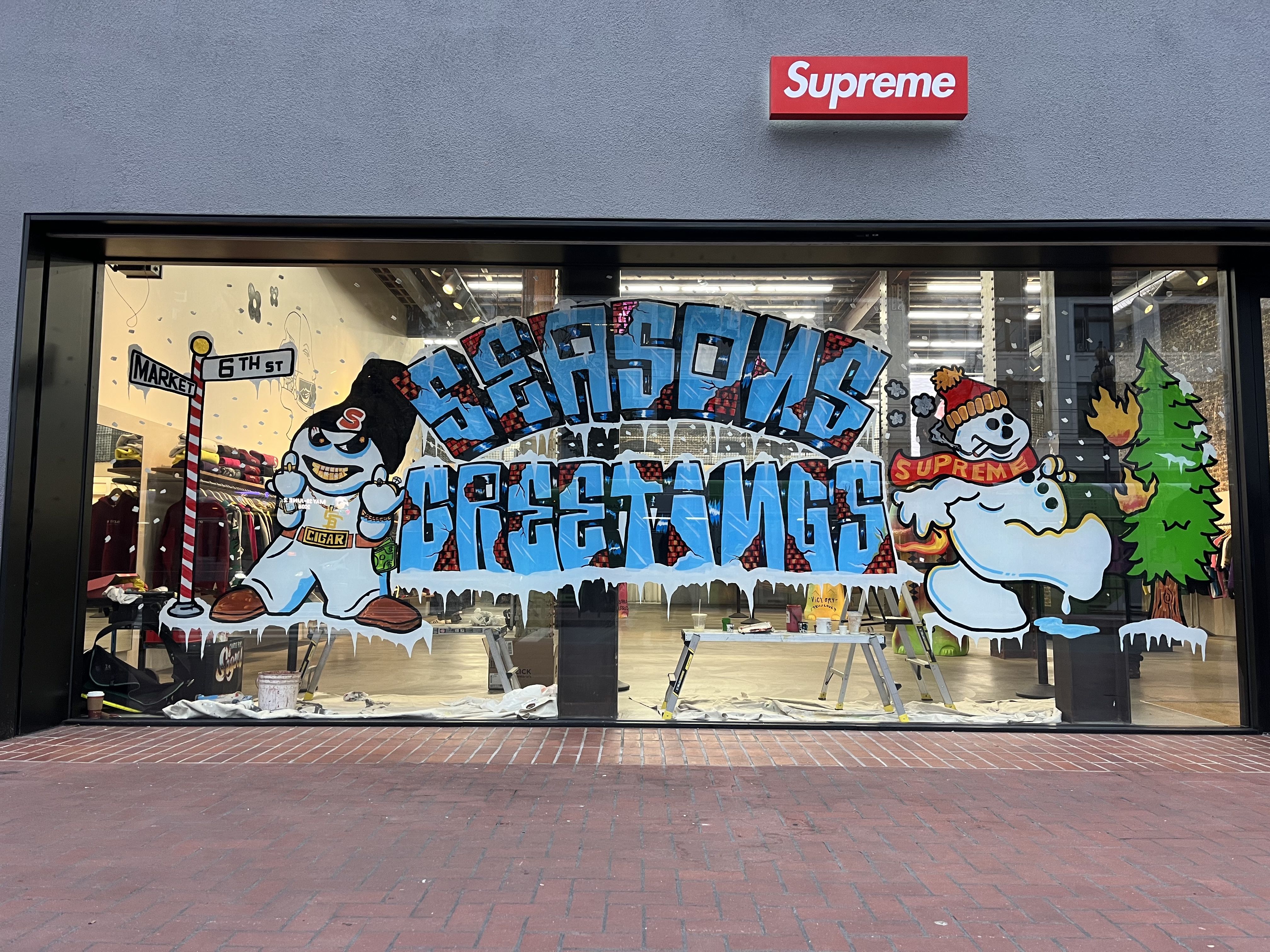 Store window with colorful graffiti spelling "SEASONS GREETINGS" in blue, flanked by two cartoon snowmen in winter attire, one near a street sign, the other next to a burning tree.