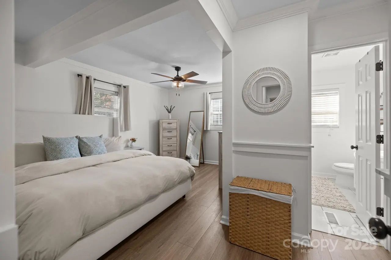Bright bedroom with beige bedding, two blue patterned pillows, ceiling fan, tall dresser, standing mirror, light wood floor, wicker basket, and partially open door to white bathroom.