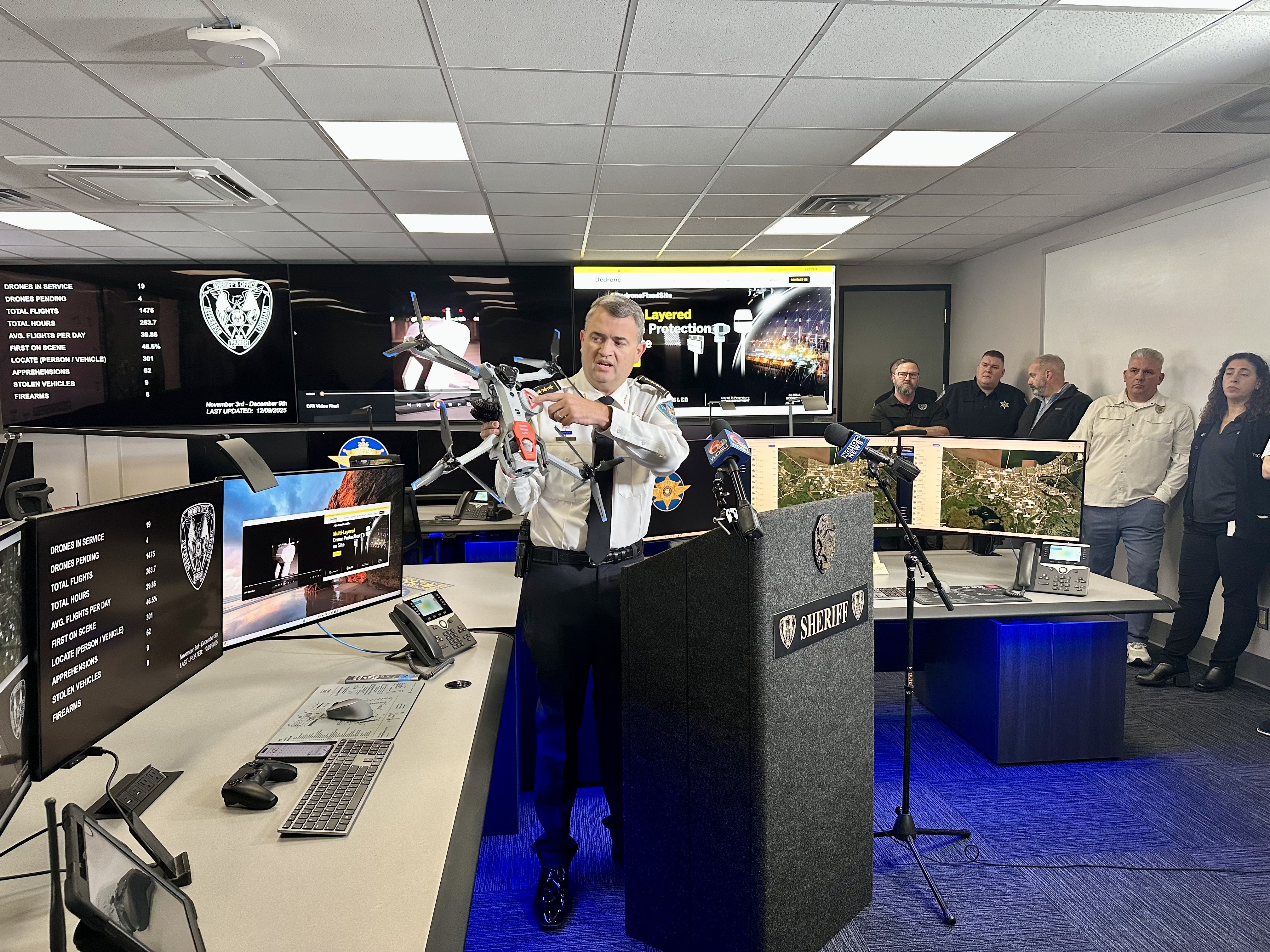 Sheriff in uniform holding a police drone, speaking at a press conference with monitors showing drone data and law enforcement stats, with several people standing behind him.