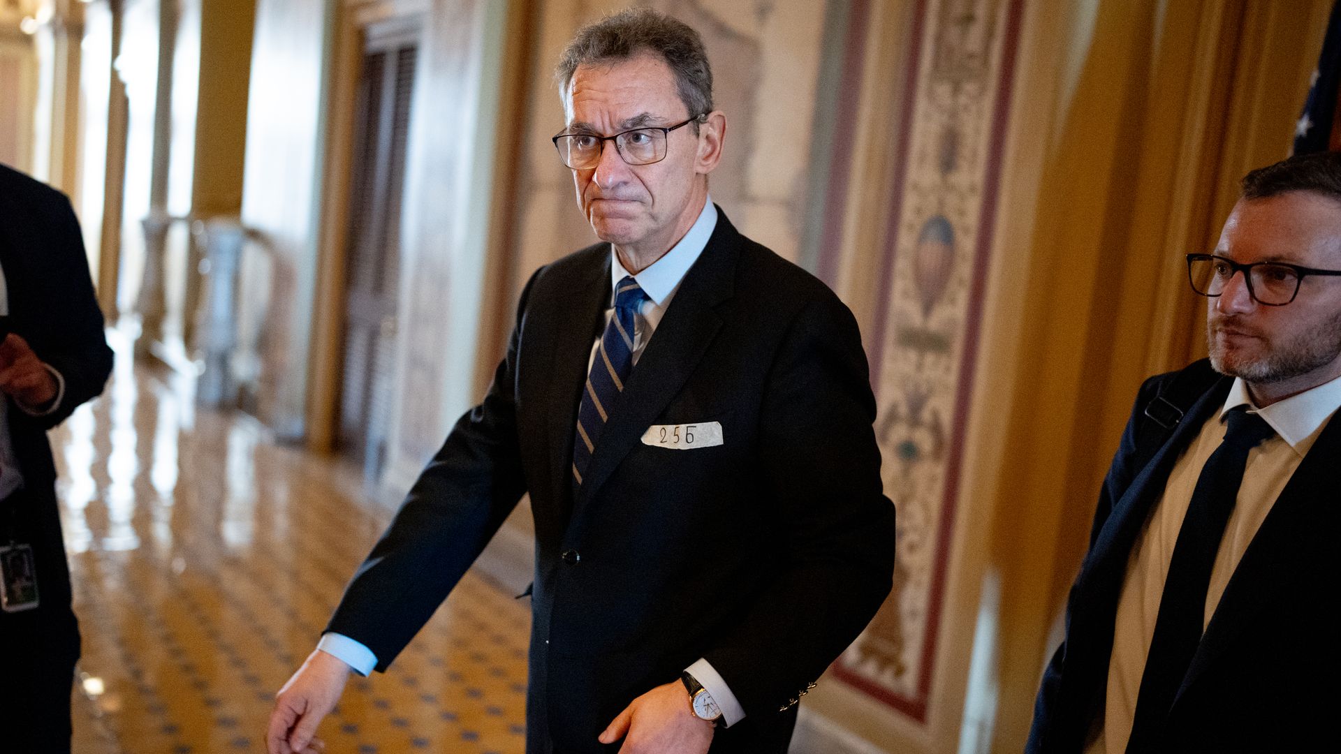A person in a black suit walks through a stately hall