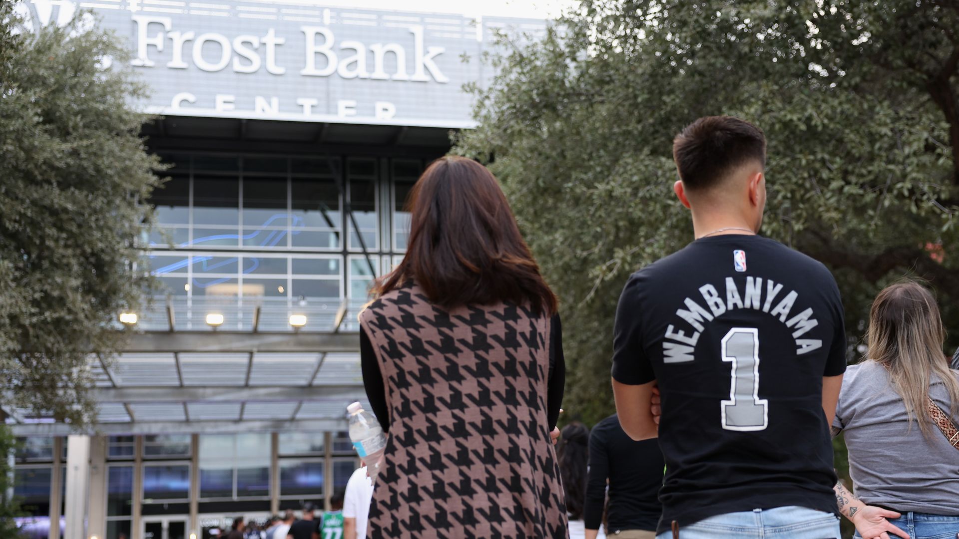 Two people stand outside in front of the Frost Bank Center entrance. One is a fan wearing a black Wembanyama shirt.