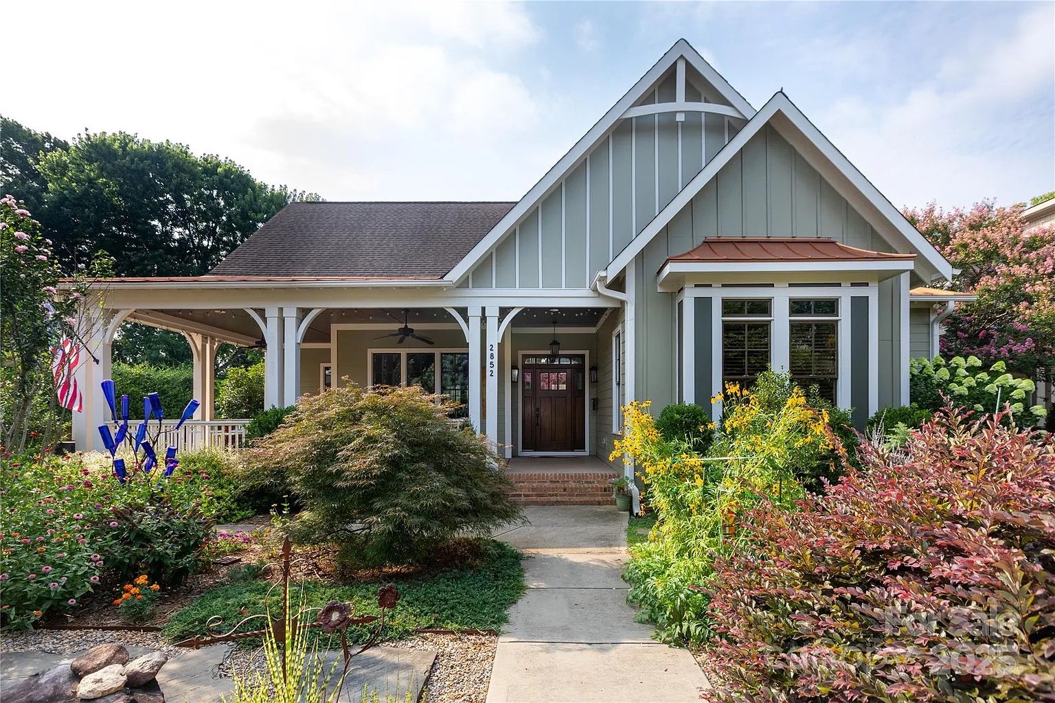 Gray house with white trim, a dark wooden door, and a covered front porch. Lush garden with green, yellow, red plants, and blue glass decorations line the stone pathway.