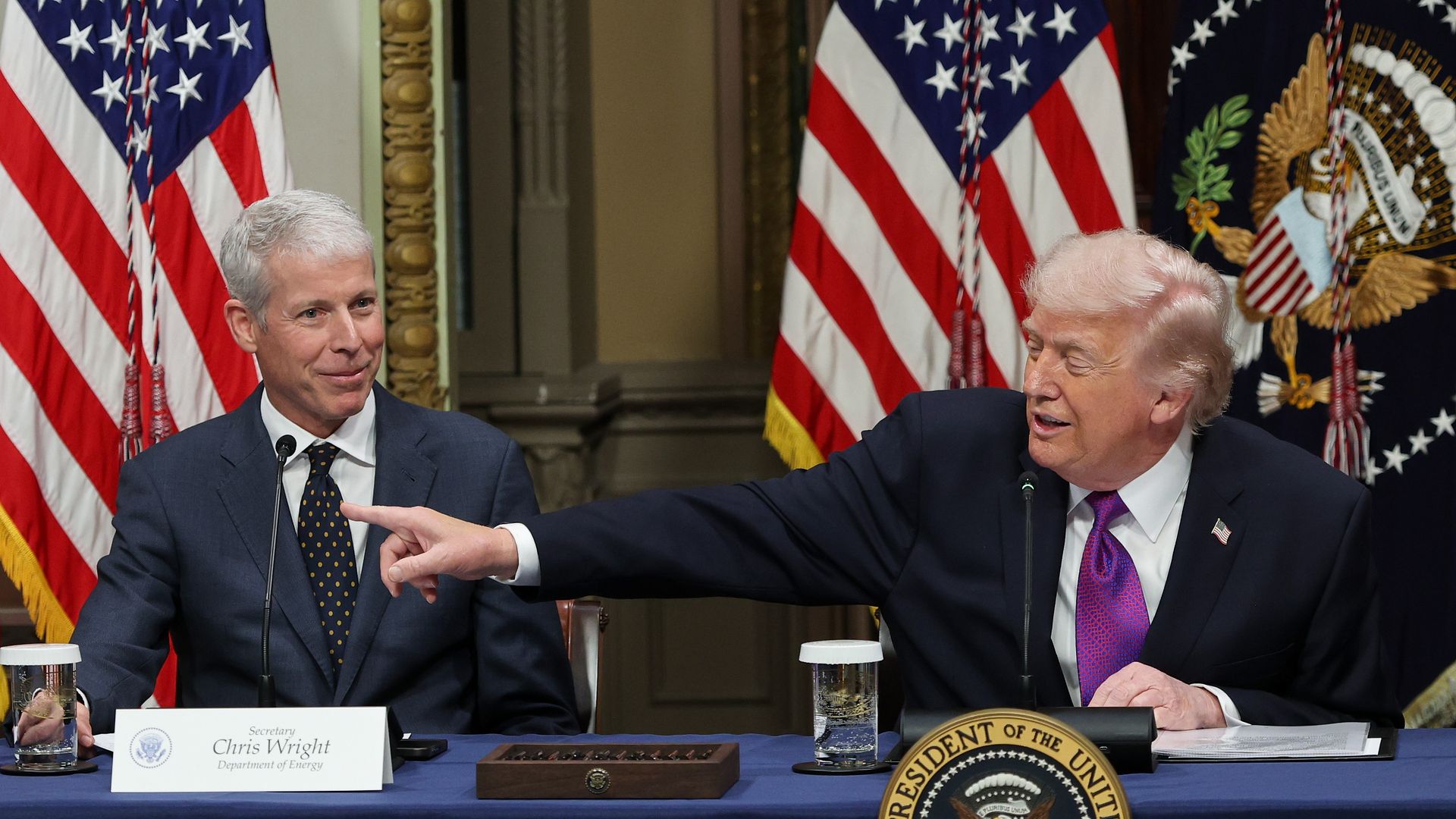 President Trump — wearing a black suit, a white collared shirt, a purple tie and an American flag pin — points as he sits at a table next to Energy Secretary Chris Wright, who is wearing a gray suit, a black and yellow polka-dot tie and a white collared shirt.