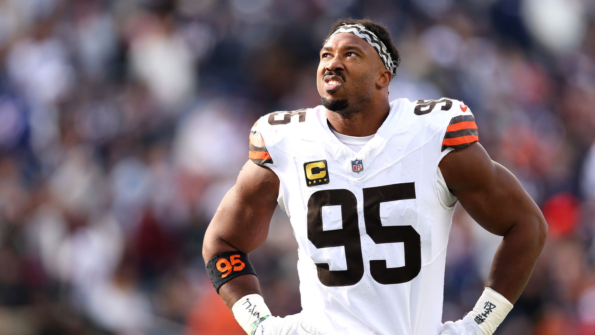 A Black football player in a white jersey with the number 95 and a captain patch, wearing a black-and-white patterned headband, gazes upward with a determined expression on the field, blurred stadium crowd in background.