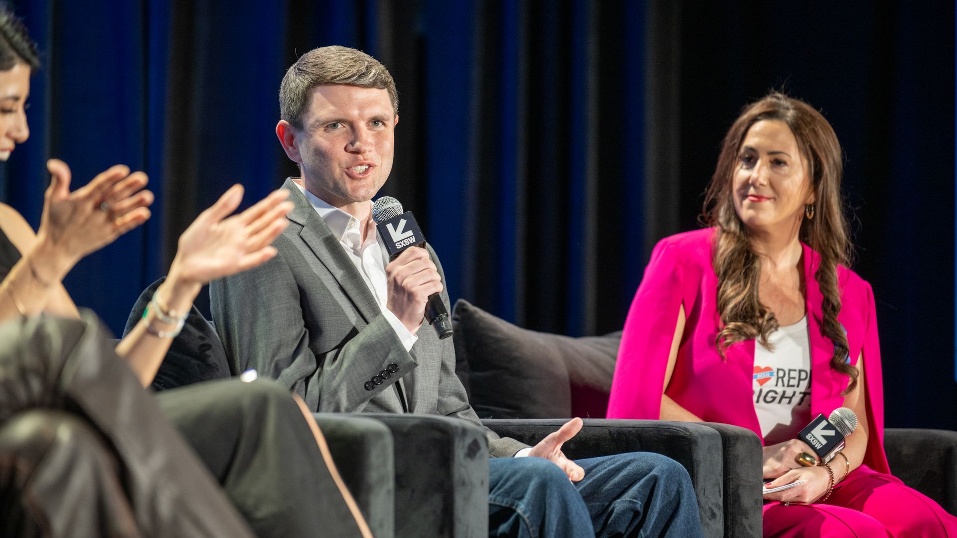 A person seated talks into a microphone, while people clap and listen.