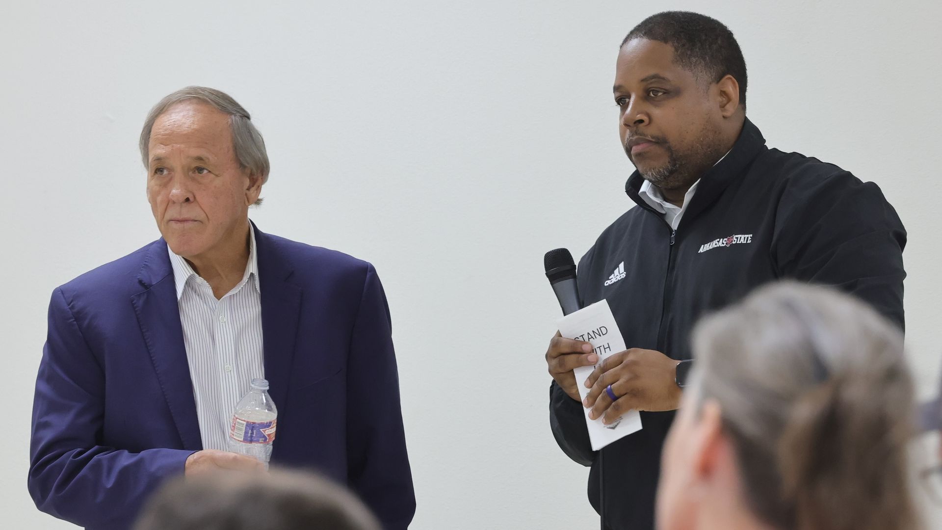 Two men stand in front of a white wall speaking to an audience. One, wearing a blue suit and holding a water bottle, looks ahead. The other, wearing a black Arkansas State jacket, holds a microphone and a folded paper labeled "STAND WITH." The heads of audience members are visible in the foreground.
