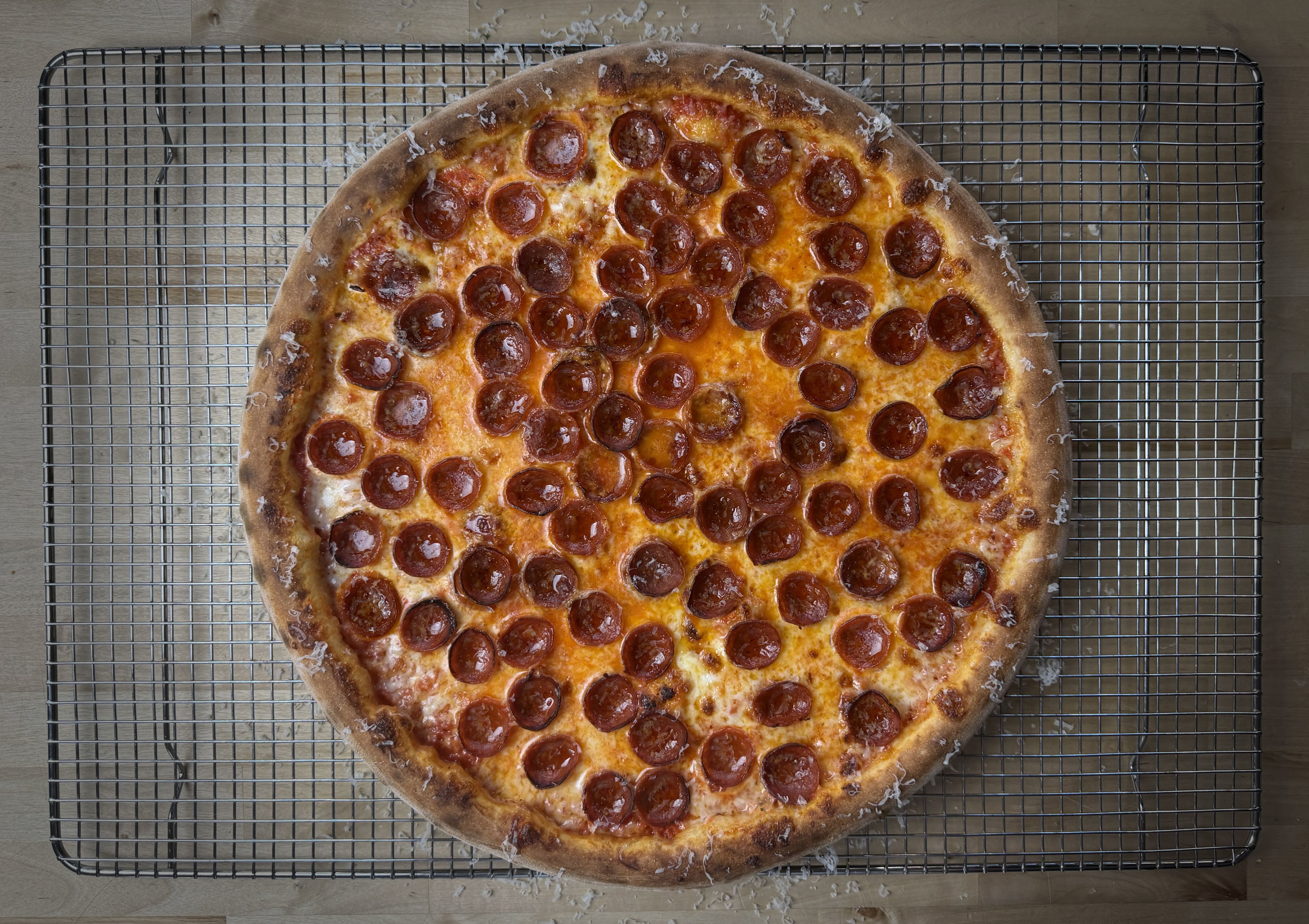Top-down view of a large pepperoni pizza cooling on a metal rack over a wooden table; bubbly cheese, golden crust, and abundant pepperoni slices.