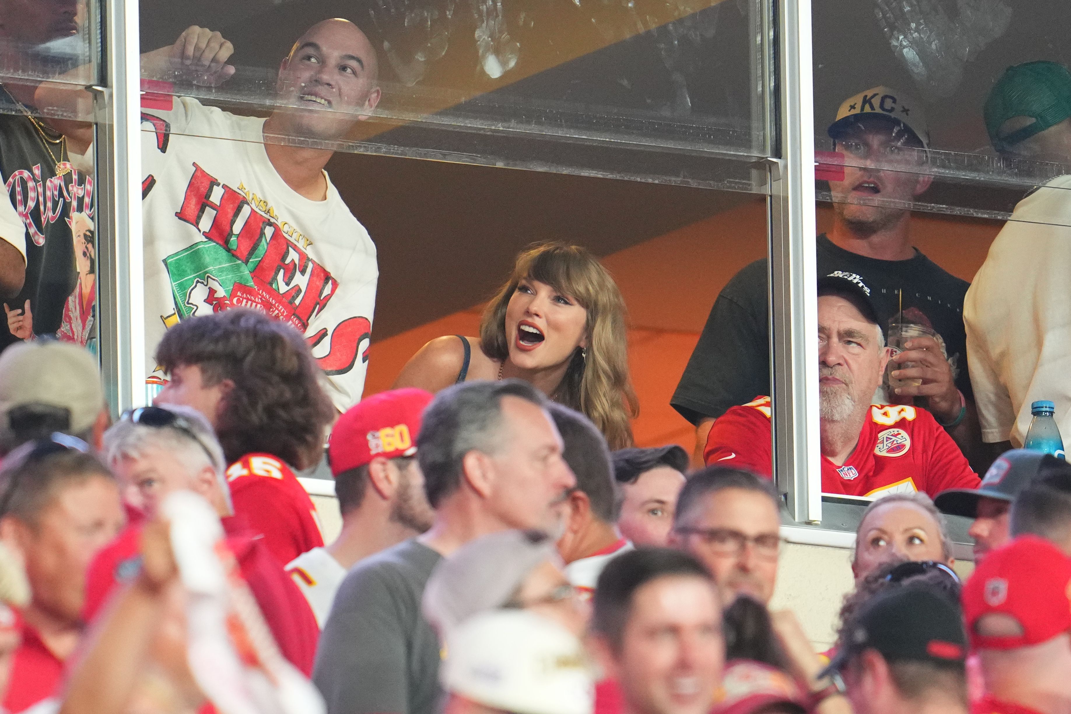Taylor Swift, standing in a suite in Arrowhead Stadium, leans down to look out a window.
