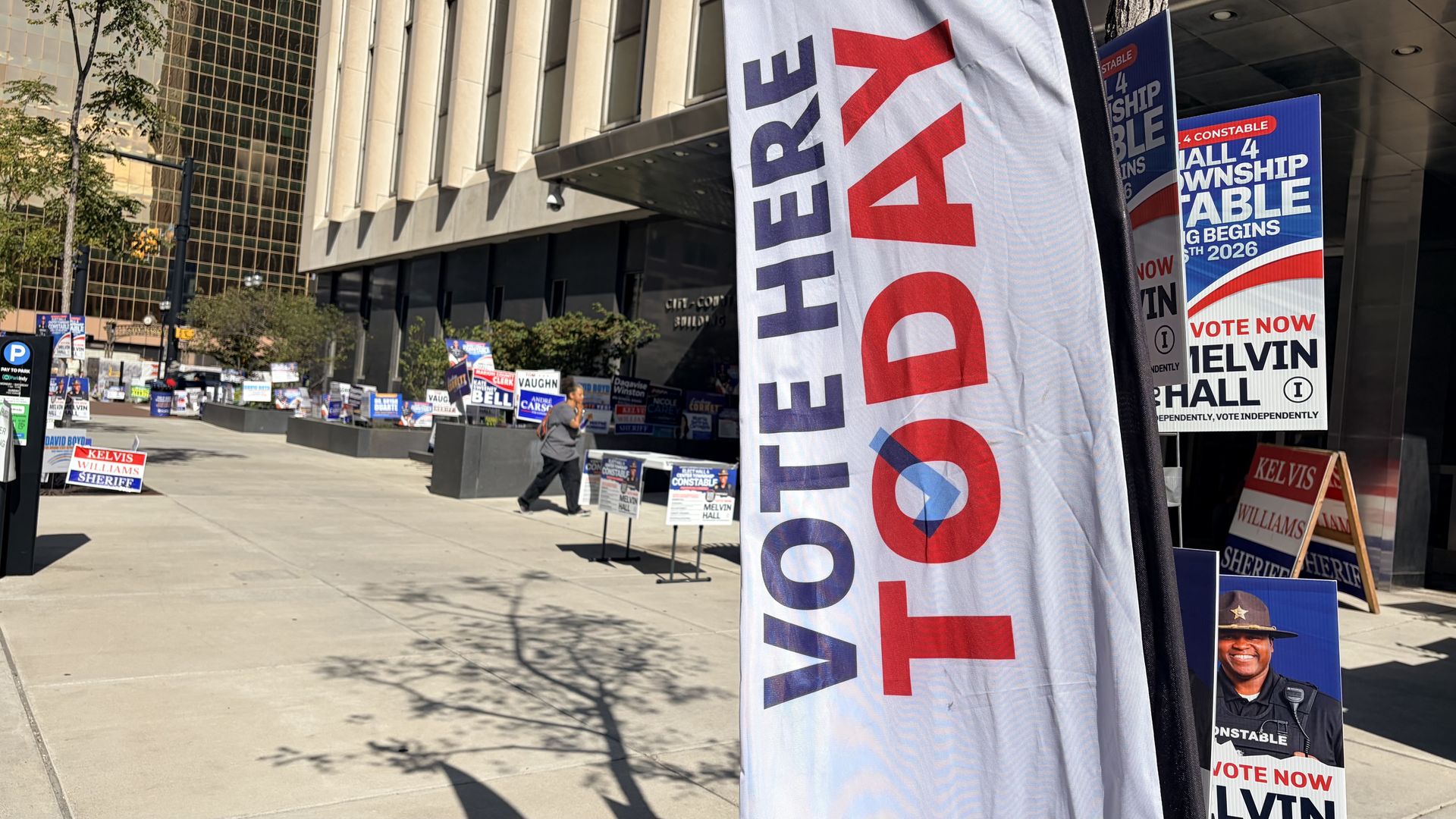 Urban sidewalk lined with red, white, and blue campaign signs. A tall vertical banner says VOTE on the right as a passerby walks past a modern office building with glass windows.