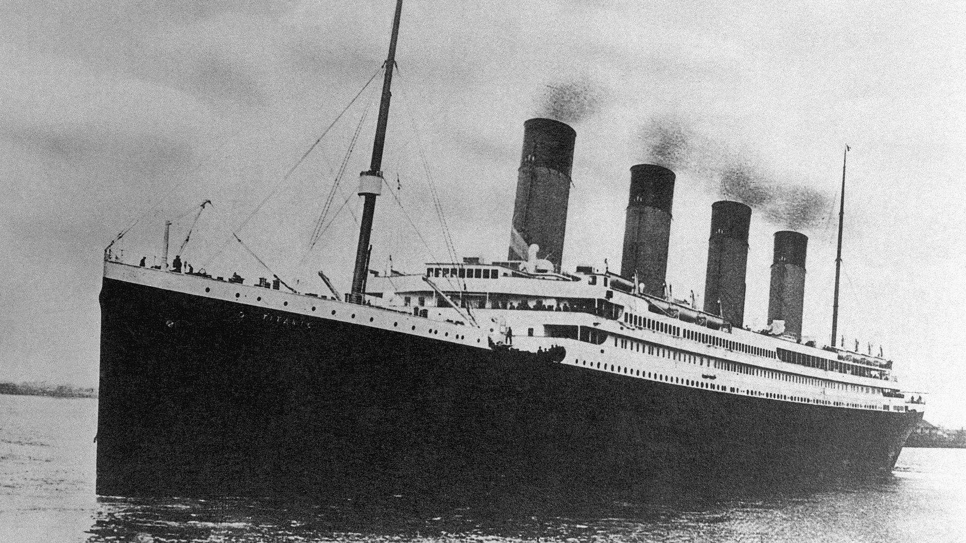 Black-and-white image of a grand early-20th-century ocean liner with four tall smokestacks, smoke rising from the funnels, white superstructure, dark hull, and passengers on the bow deck.