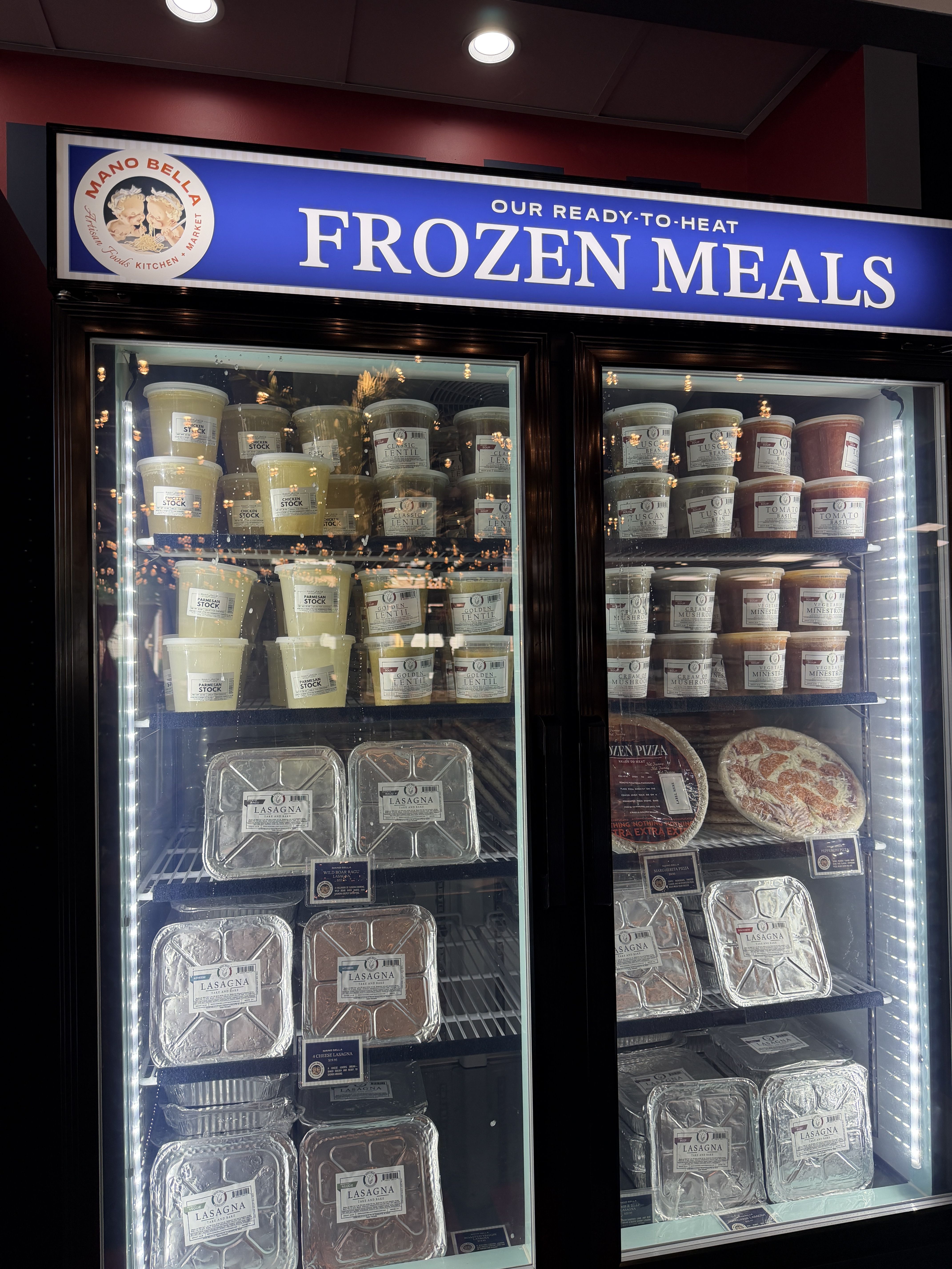 Refrigerated display with a blue sign reading "OUR READY-TO-HEAT FROZEN MEALS". Left logo circle; shelves show stacked plastic containers and lasagna trays behind glass.