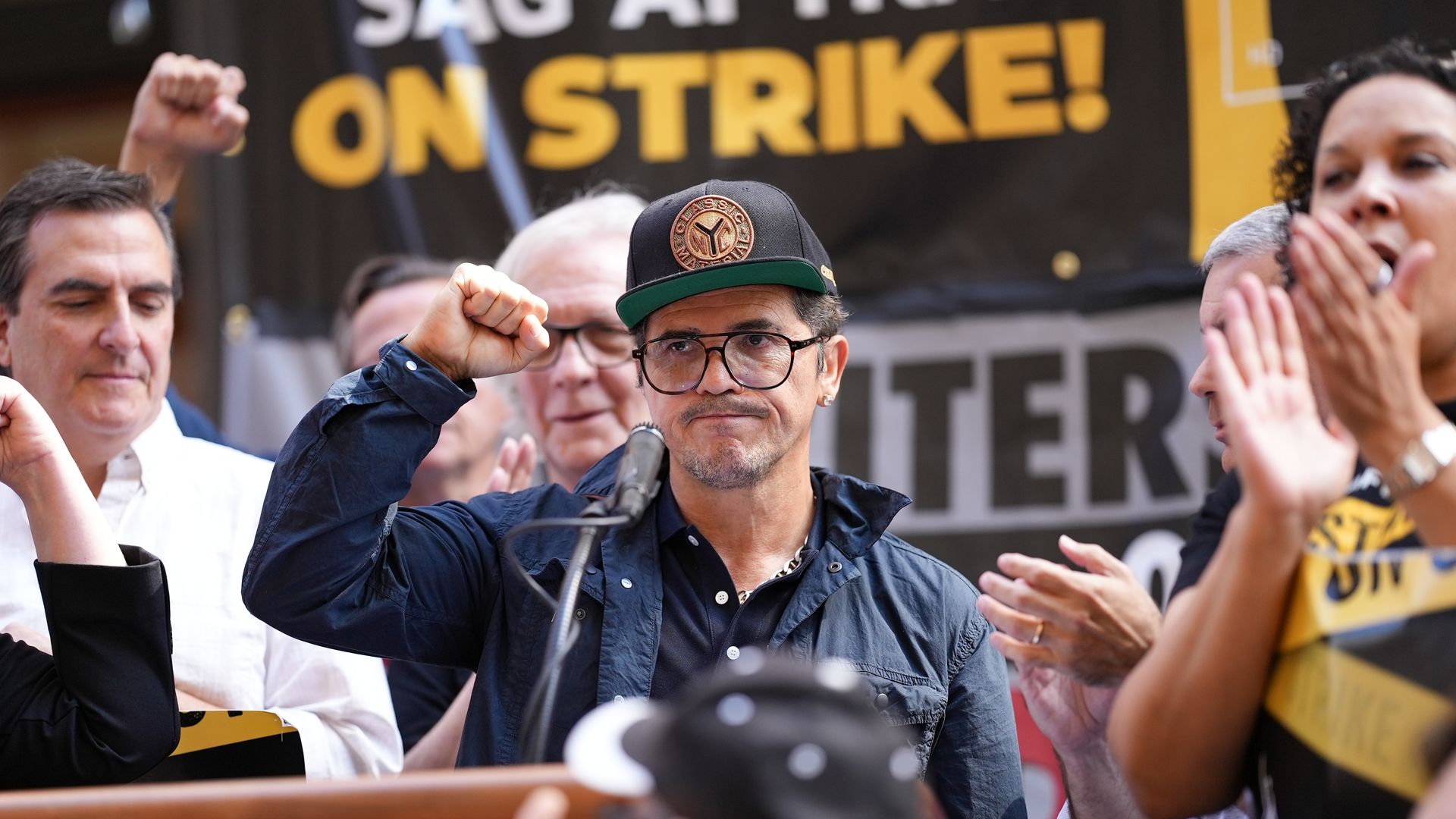 Actor John Leguizamo, standing at a microphone podium with other people, raises his fist in the air during a press conference for the SAG actors strike