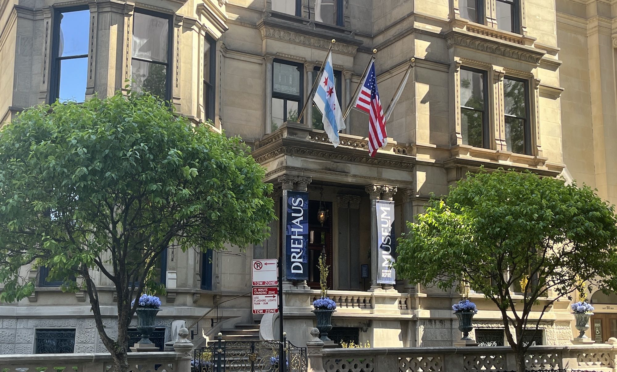 Historic stone building with ornate façade. United States and Chicago flags fly from a balcony; blue banners read "MUSEUM" beside the entrance, with green trees in the foreground.