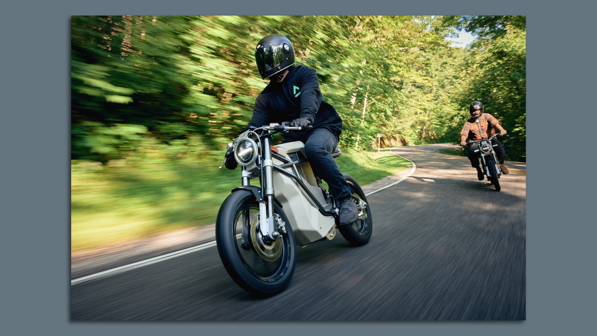 A photo of two people riding Land electric motorcycles around a bend in a sunny, forest-lined road.