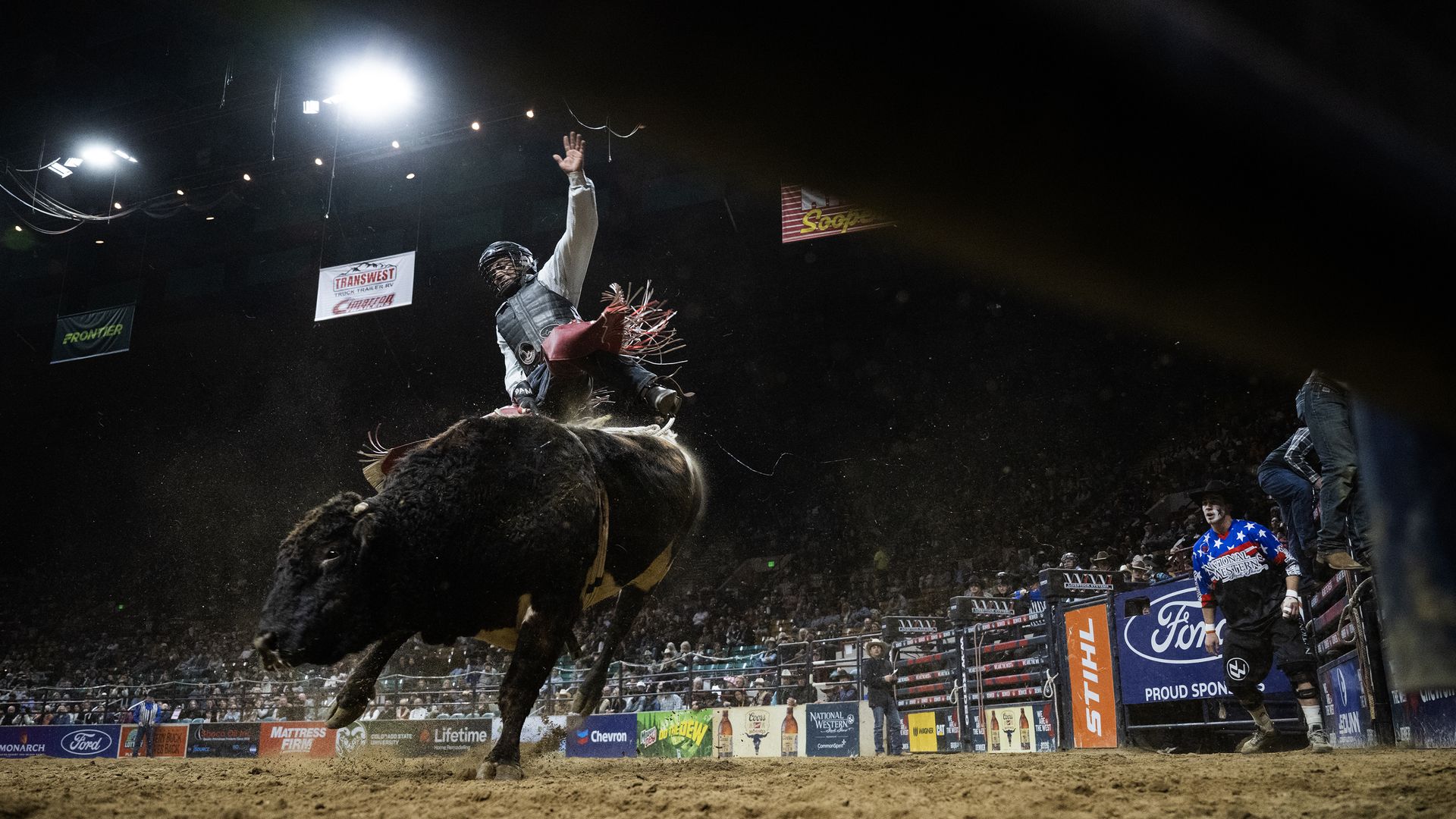 A cowboy wearing a protective vest and helmet rides a bucking bull in a dim arena under bright overhead lights, raising one hand as banners line the arena and a cheering crowd watches.