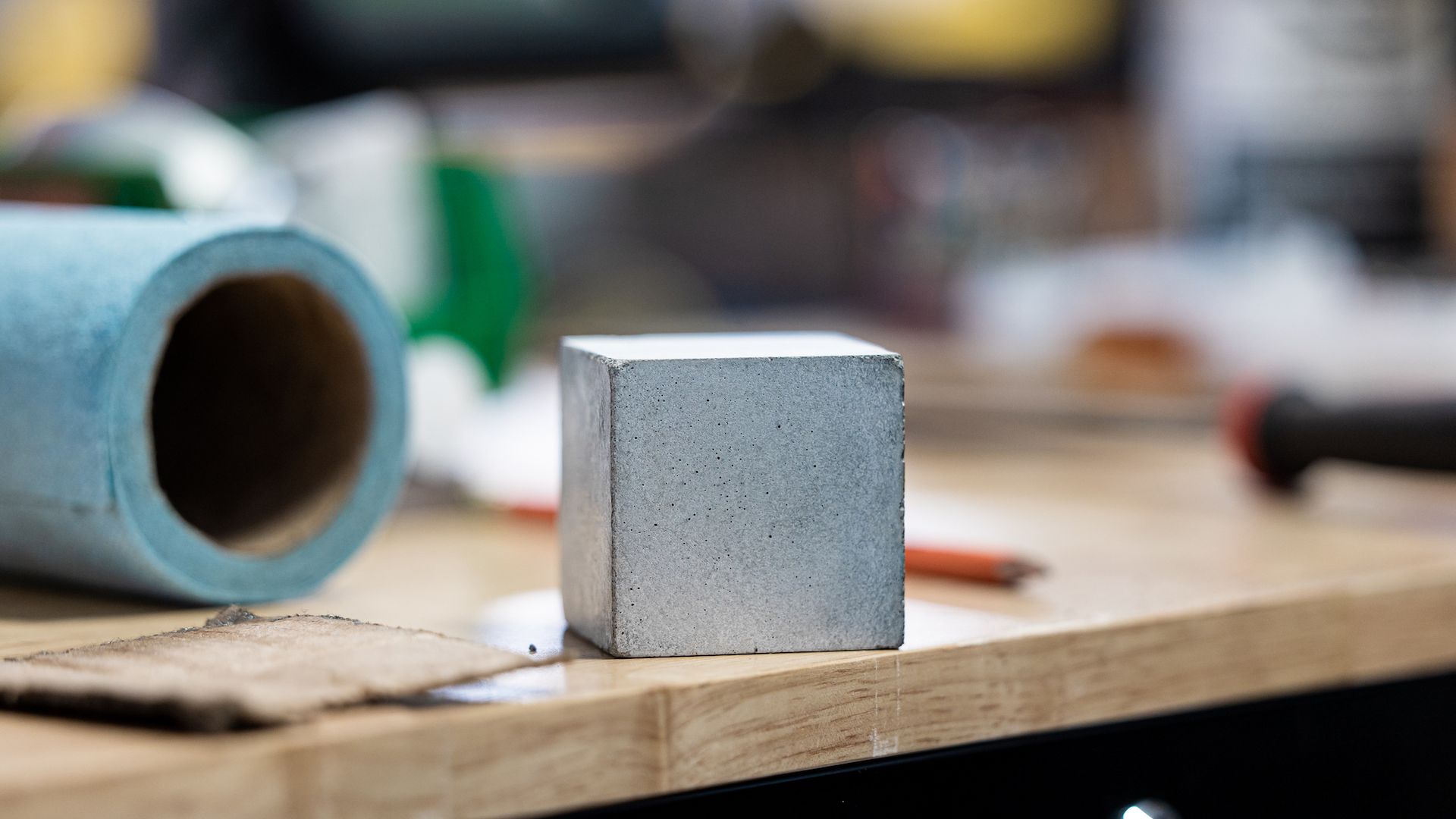 Gray concrete cube on a wooden workbench with a blue roll and tools blurred in the background in a workshop setting.