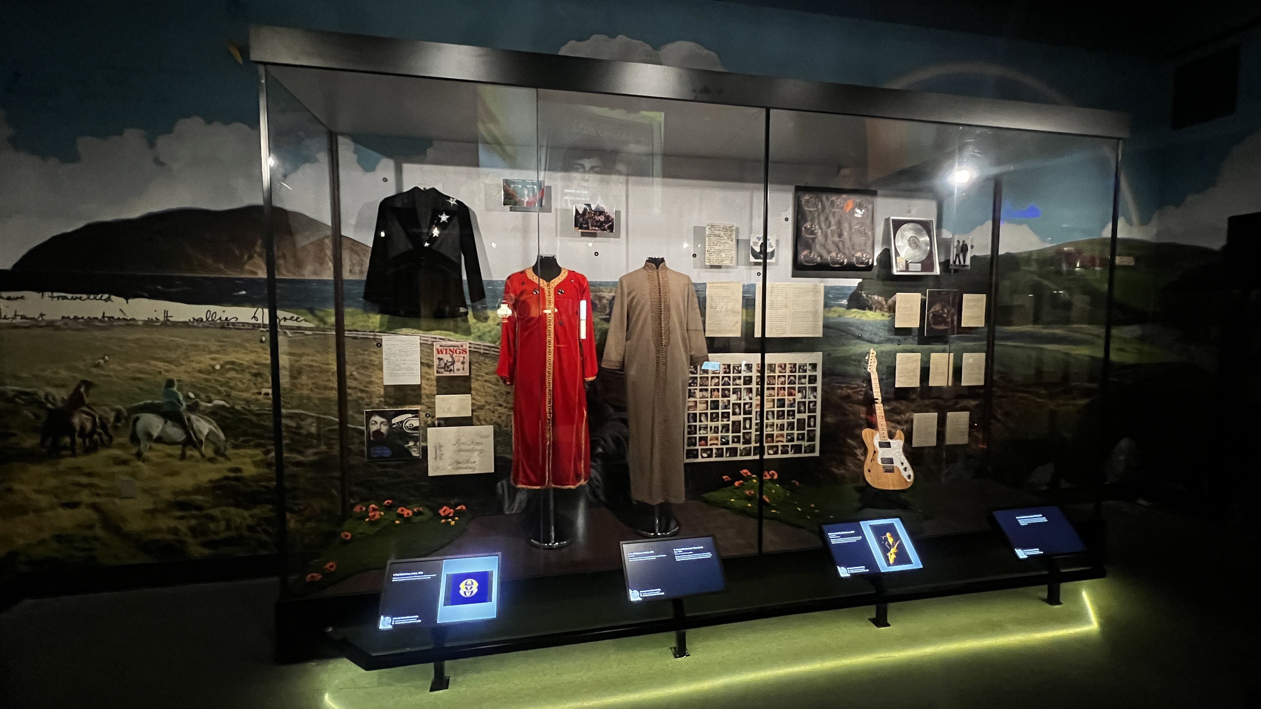 Display case with antique clothing: a black jacket with star insignias, a bright red ceremonial robe, a brown/beige long coat, a guitar, photos and documents; mural of hills and lake behind, museum lighting.