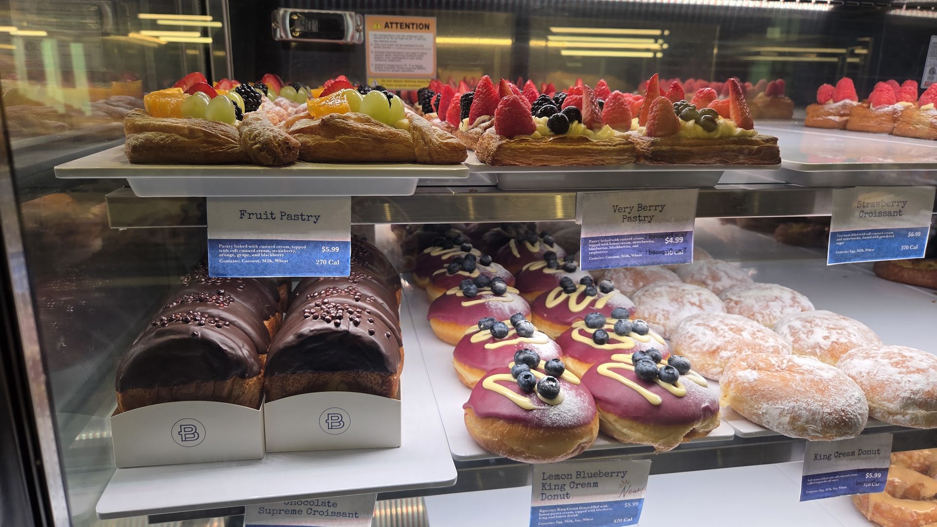 Bakery display case featuring fruit-topped croissant pastries with custard, grapes and berries; lower shelf shows chocolate and pink-frosted donuts with blueberries.