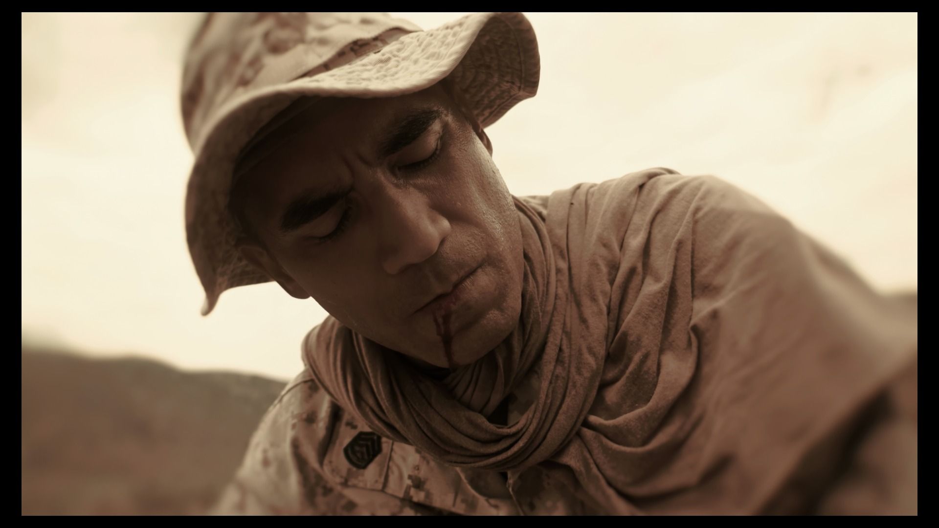 Sepia-toned close-up of a wounded soldier in desert fatigues and scarf, eyes closed, blood at his lip, head bowed as a barren landscape blurs in the background.