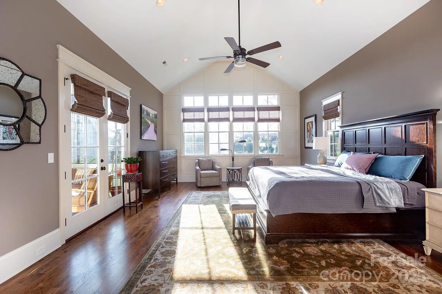 Bright bedroom with wooden floors, large windows with brown blinds, dark wood bed with blue and pink pillows, two armchairs near the window, a ceiling fan, and a decorative rug.