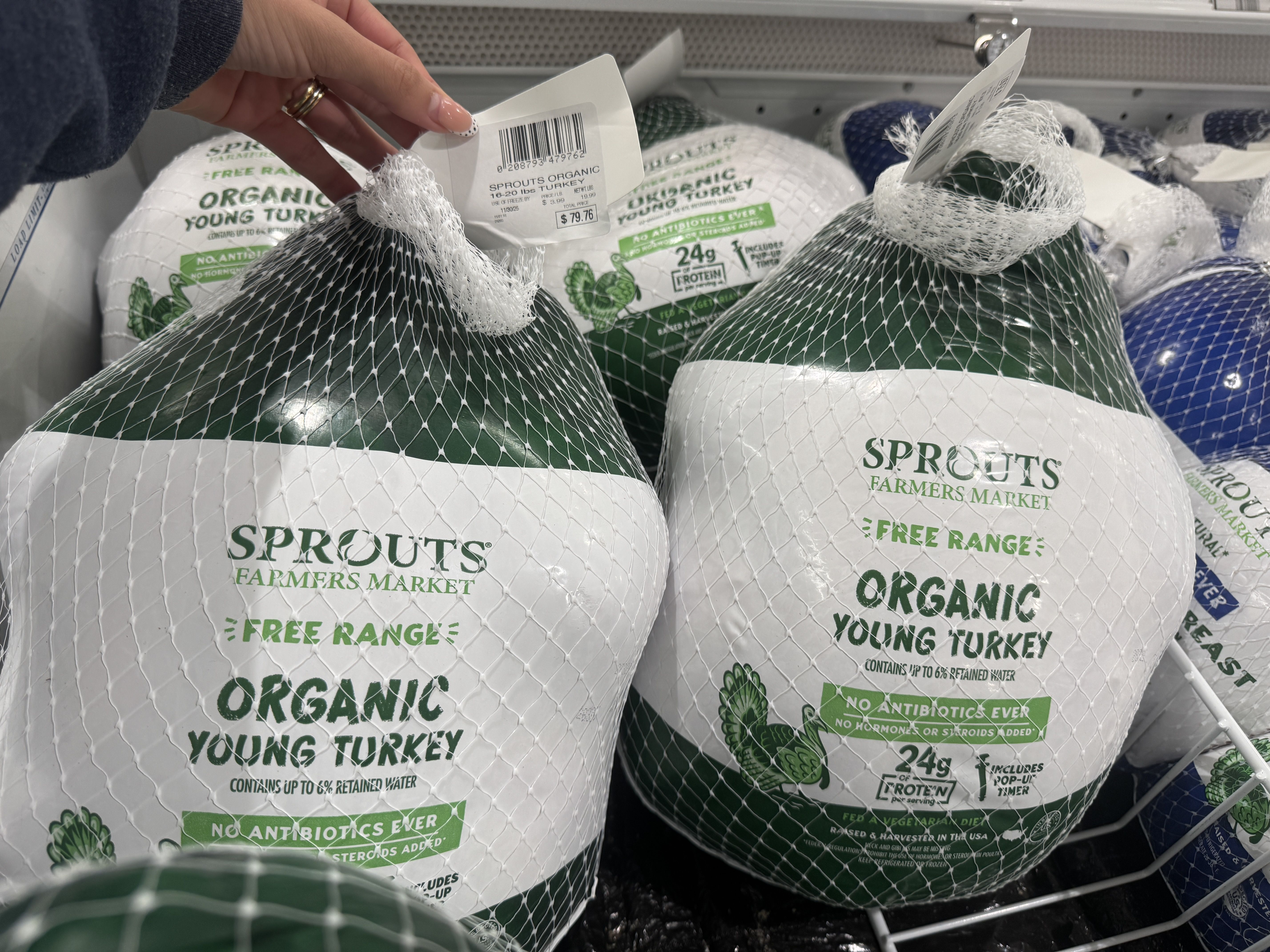 Hand holding a price tag on a netted green and white packaged organic young turkey from Sprouts Farmers Market labeled free range, no antibiotics, 24g protein, priced $79.76.