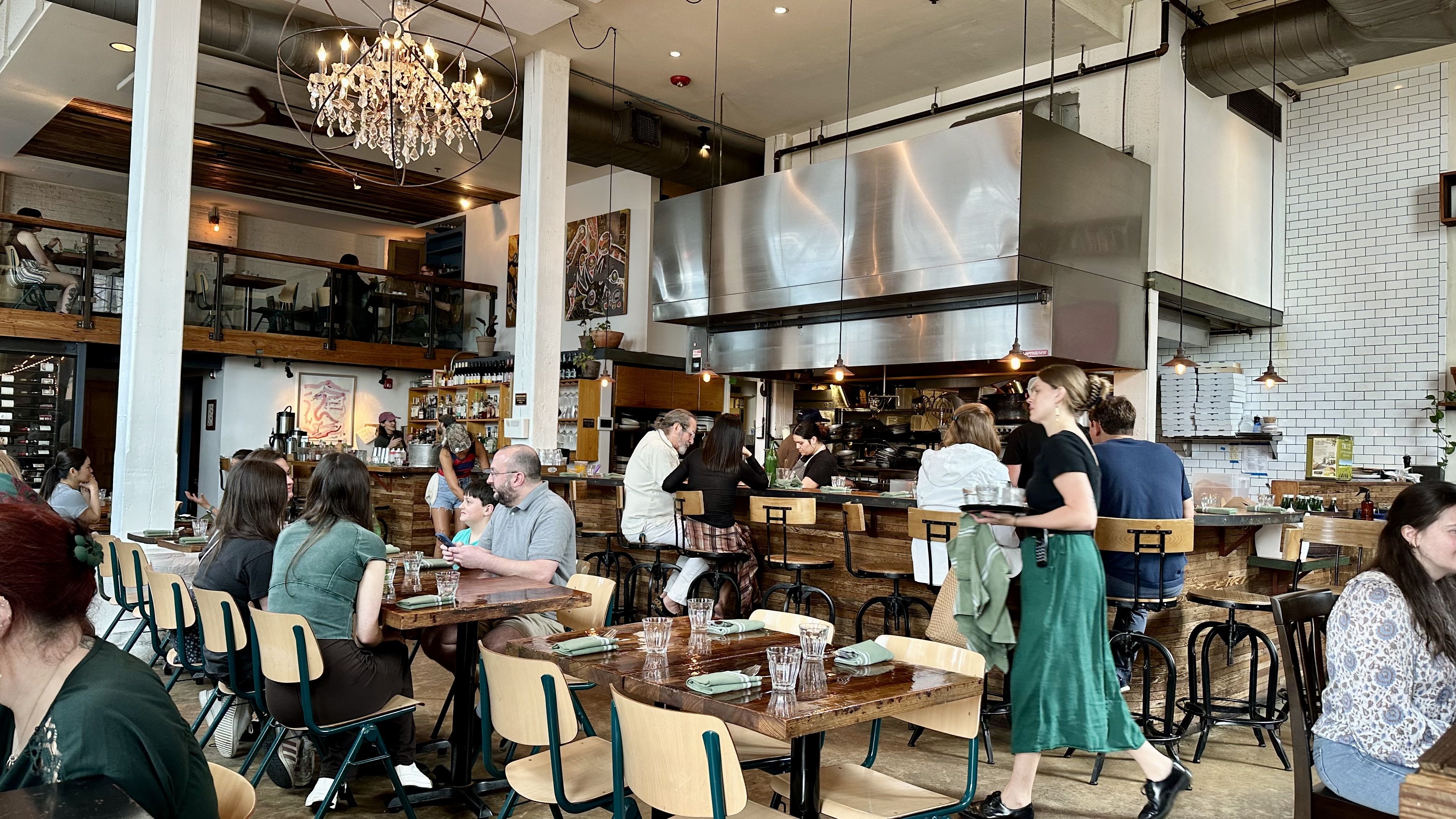 Busy cafe interior with an open stainless-steel kitchen hood, chandelier, and hanging bulbs. Two-level seating with wooden tables and customers dining; staff serve near the kitchen.
