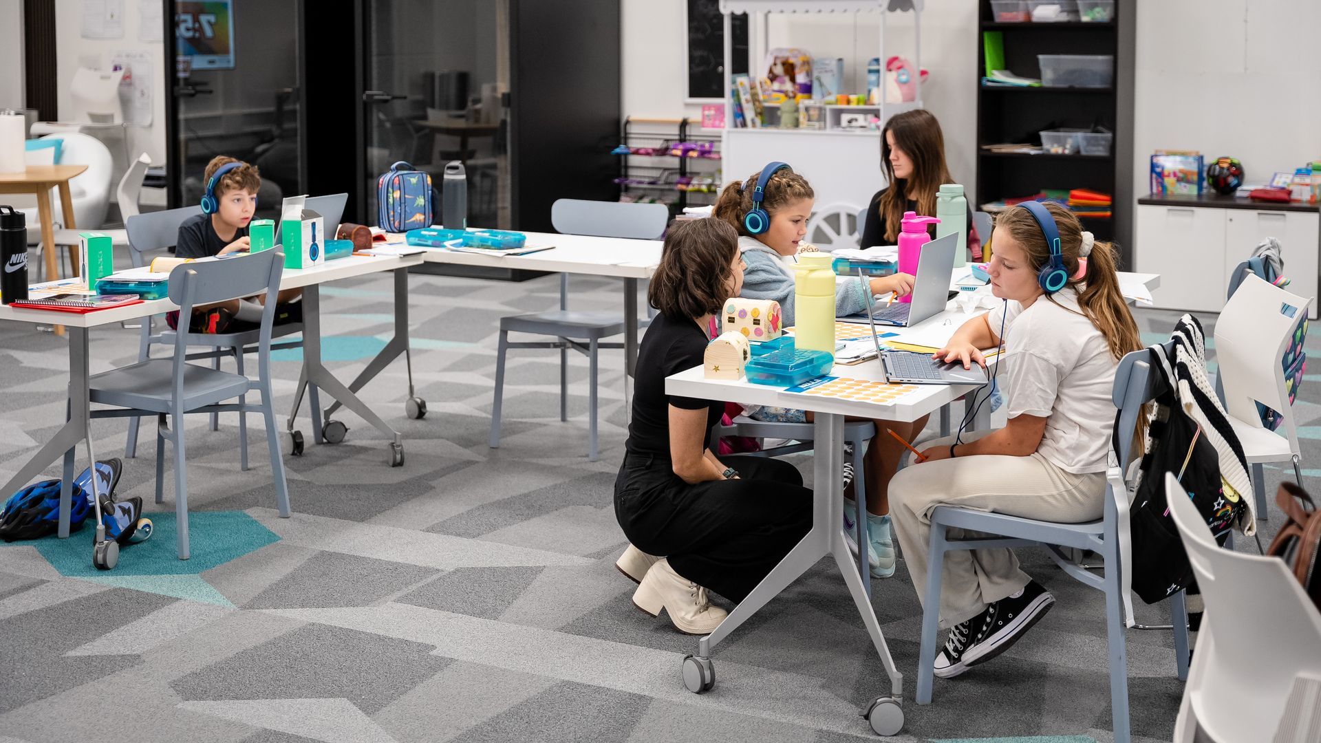 Children wearing blue headphones working on laptops at tables in a bright classroom with colorful water bottles, a teacher kneeling to assist, and shelves with supplies in the background.