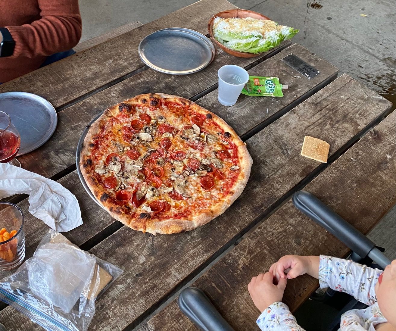 A pepperoni and mushroom pizza on a rustic wooden table, with an empty plate, a lettuce wedge salad topped with cheese, a plastic cup, a green GoGo squeeZ pouch, a cracker, and a child’s hands resting on the table.