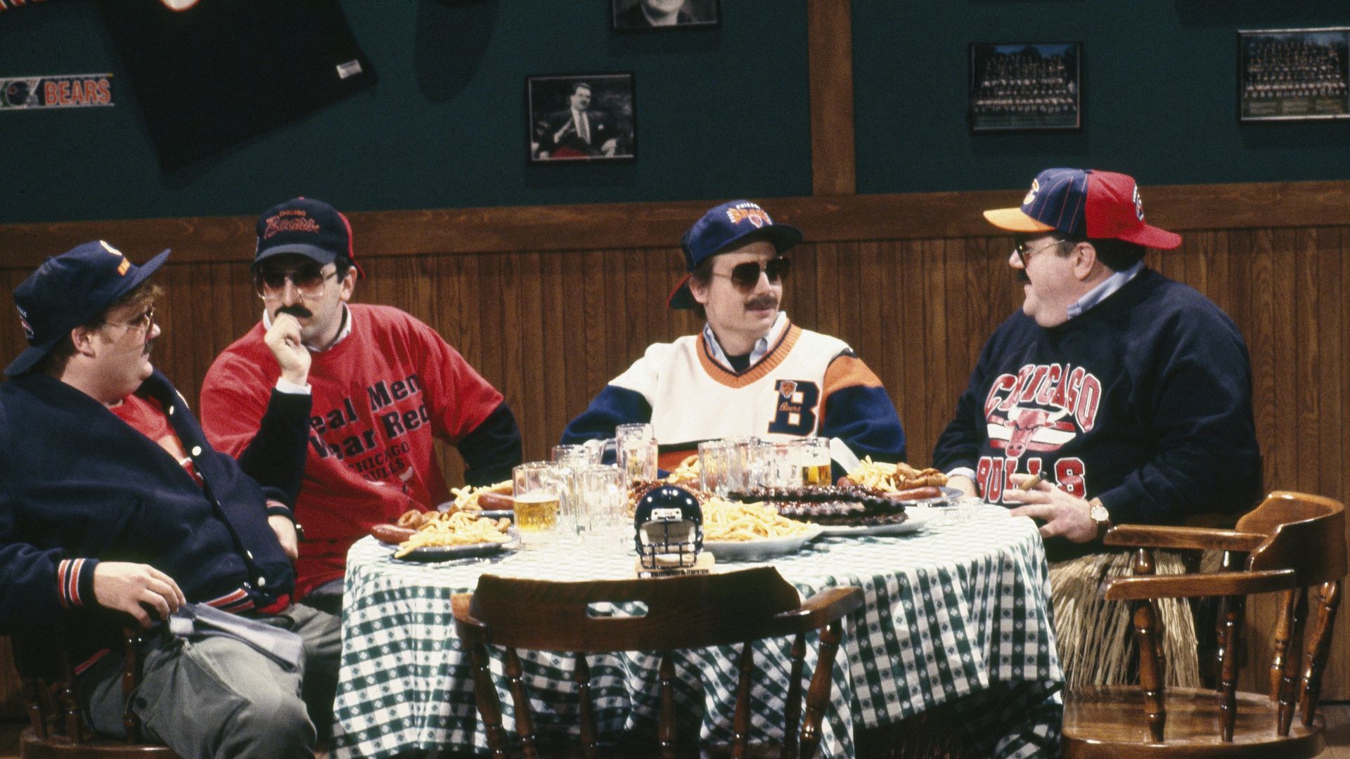 Chris Farley, Robert Smigel, Mike Myers and George Wendt sitting at a round table wearing Chicago Bears shirts and hats.