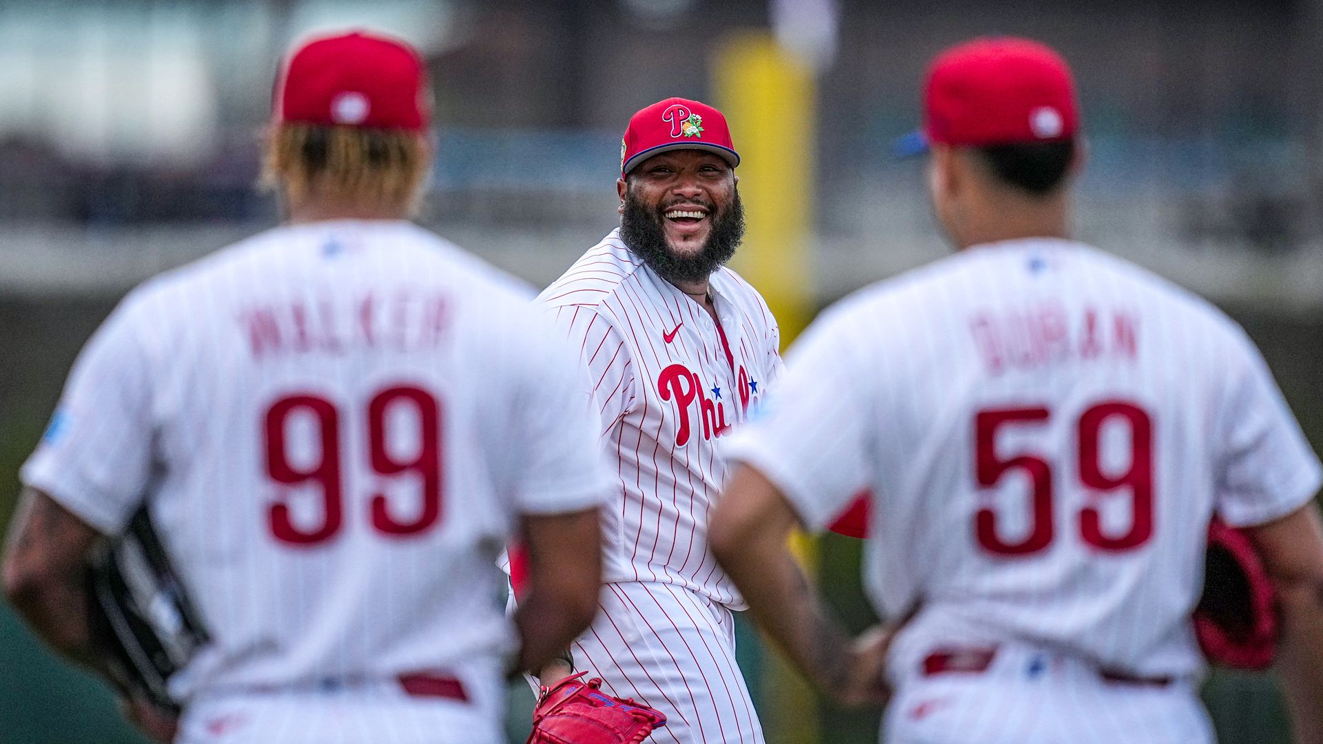 Three Philadelphia Phillies baseball players in white pinstriped uniforms and red caps; one smiling player facing forward with a red glove, two blurred players seen from behind with numbers 99 and 59.