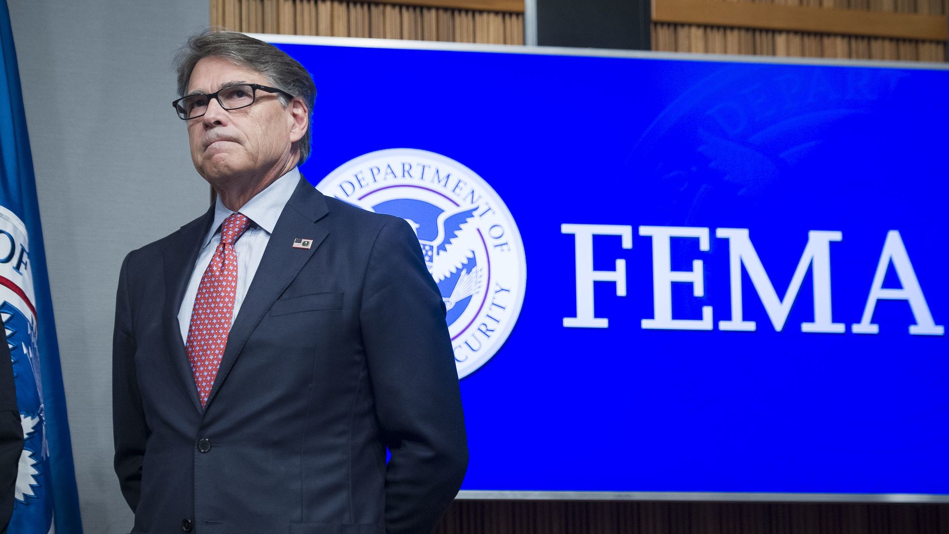 Energy Secretary Rick Perry stands with his hands behind his back in front of a blue FEMA sign. 