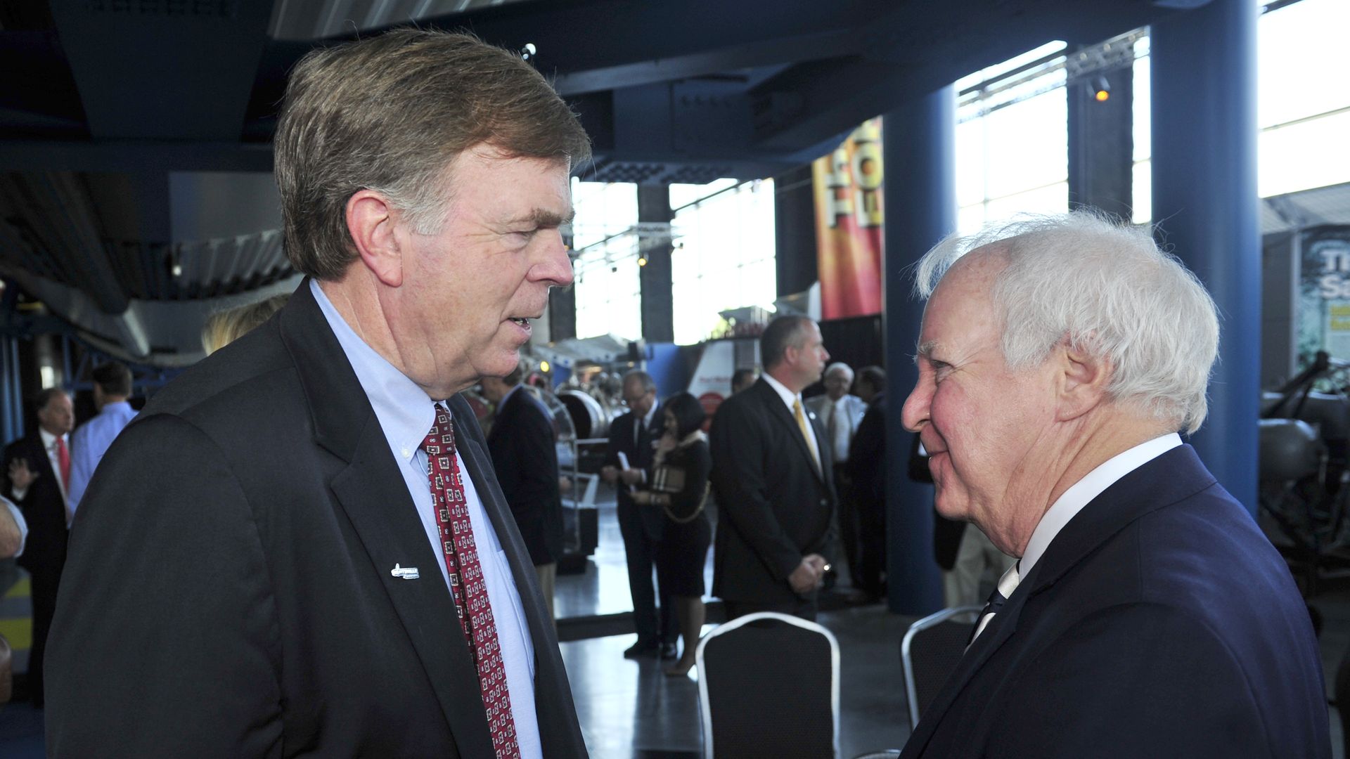 Two elderly men in suits, one with gray hair and one with white hair, engaged in conversation in a bright, modern indoor venue with people and chairs in the background.