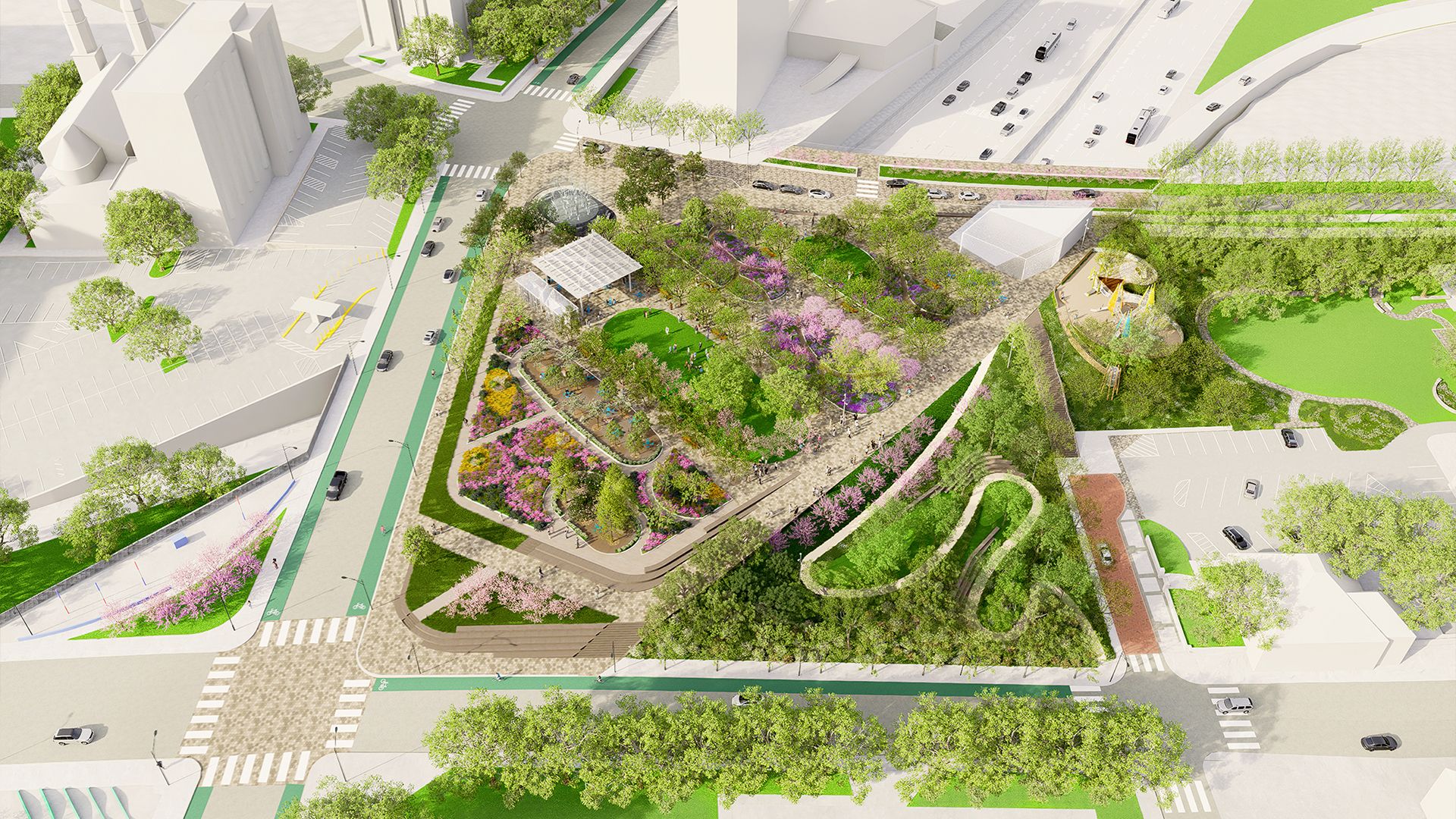 Aerial view of a triangular urban park with colorful flower beds, green lawns, walking paths, trees, a small playground, nearby streets with cars, and adjacent buildings and parking lots.