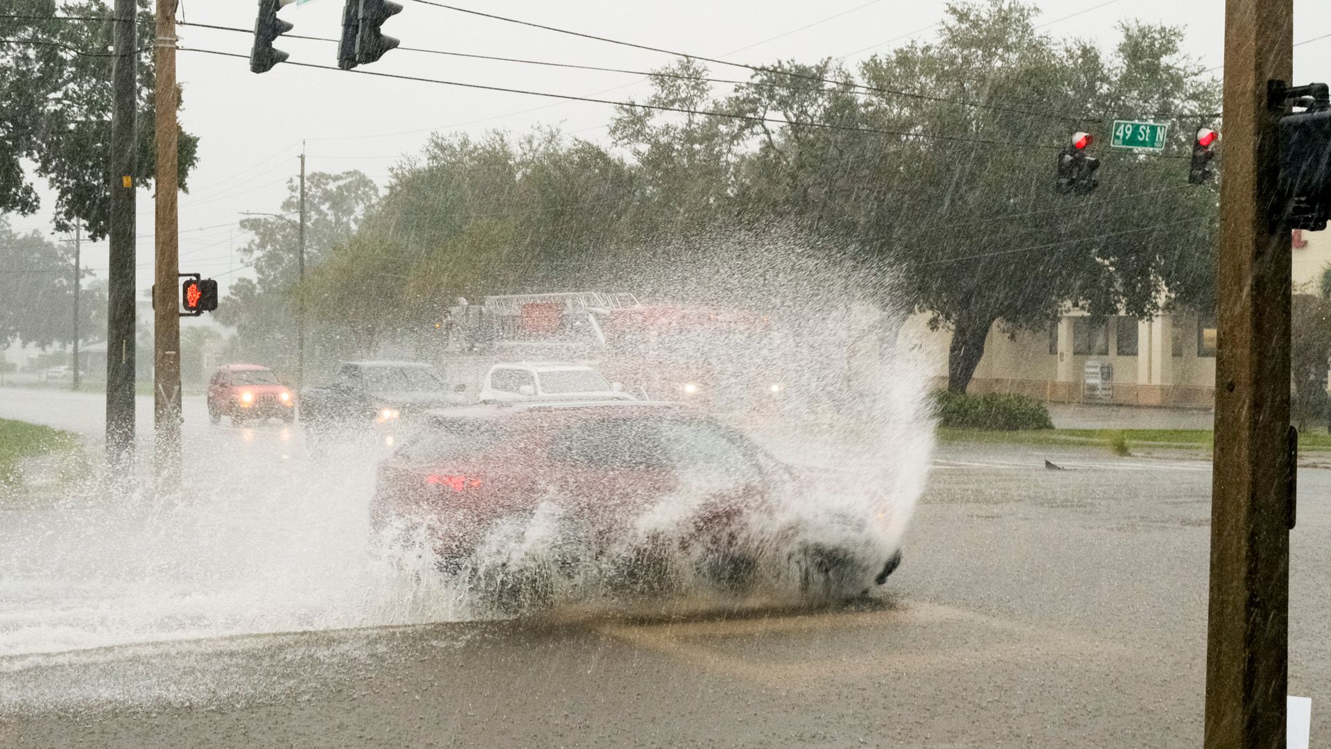 Heavy rain at a city intersection: a car creates a huge spray as it drives through floodwater; red traffic lights, green street signs (9th Ave N, 49th St N), trees and utility poles in misty background.