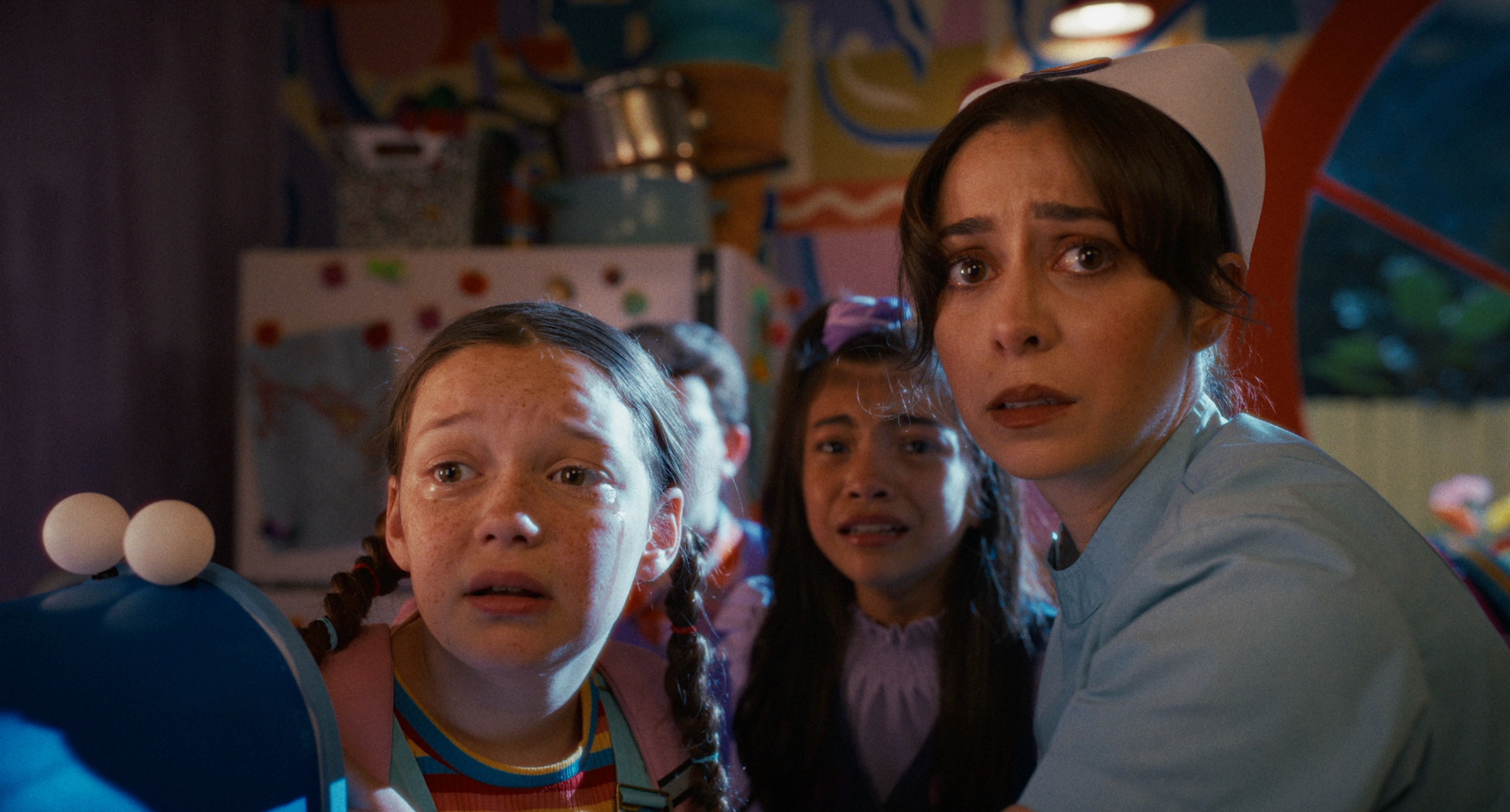 A tense scene in a colorful room: a nurse in a light blue uniform with a white cap on the right, looking anxious. In the foreground and background, children stare worriedly amid bright murals.