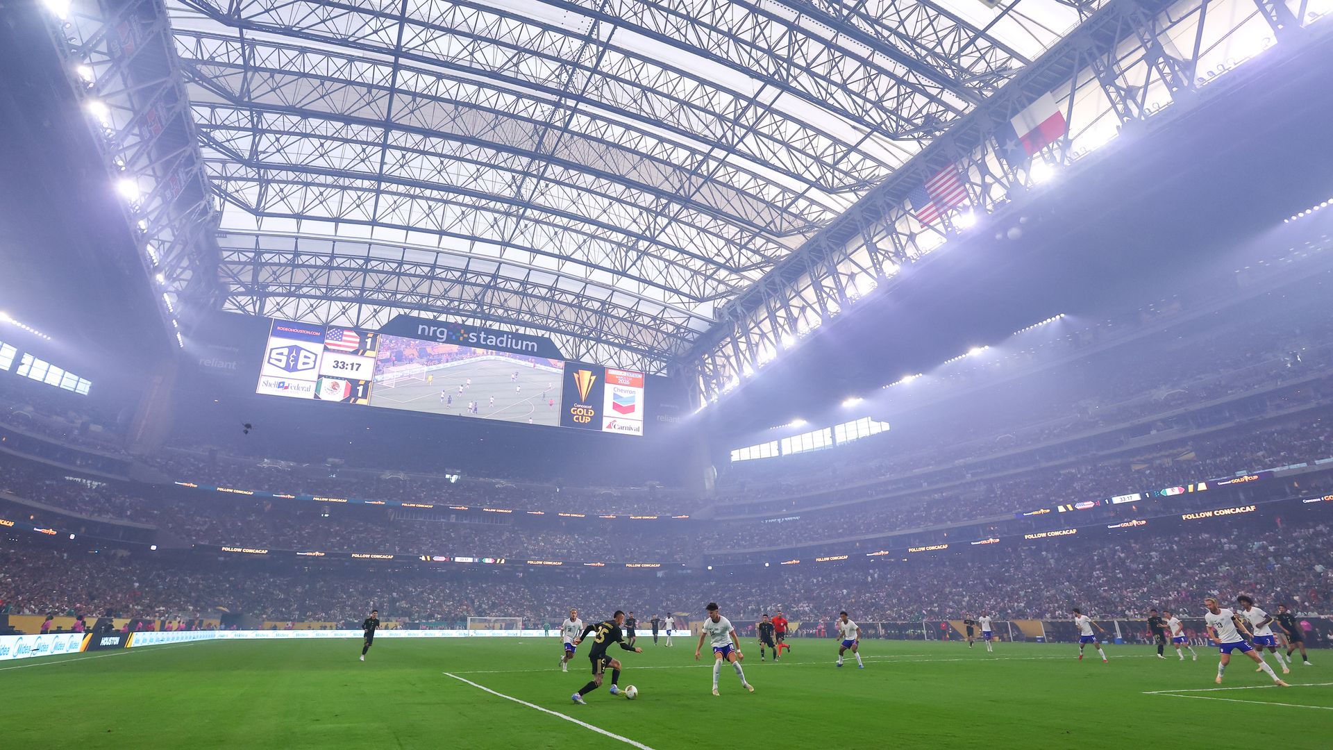 Inside a vast indoor stadium with a blue-toned steel lattice roof, flags (France and USA) hang overhead as a soccer match unfolds on a bright green pitch, with a large scoreboard and cheering crowd.